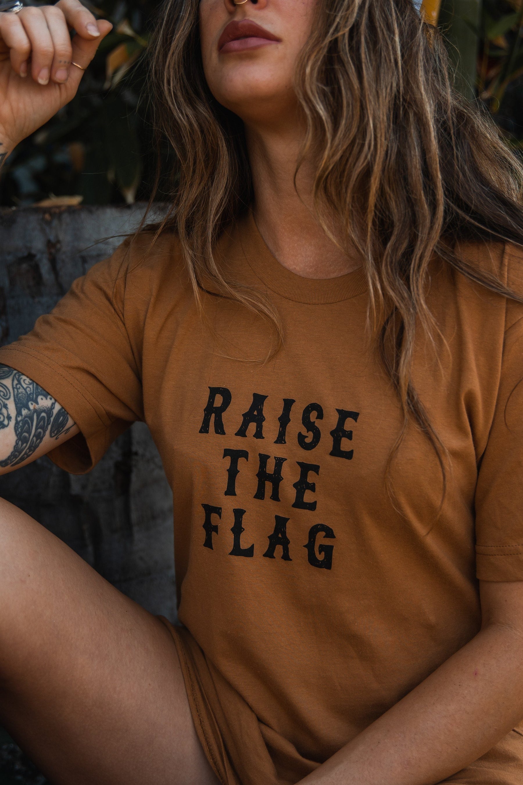 A long-haired individual sits amid lush greenery wearing Relentless Betrayals brown Raise the Flag Tee, evoking adventure and the call to either surrender or face an attack, with a tattoo on their arm.