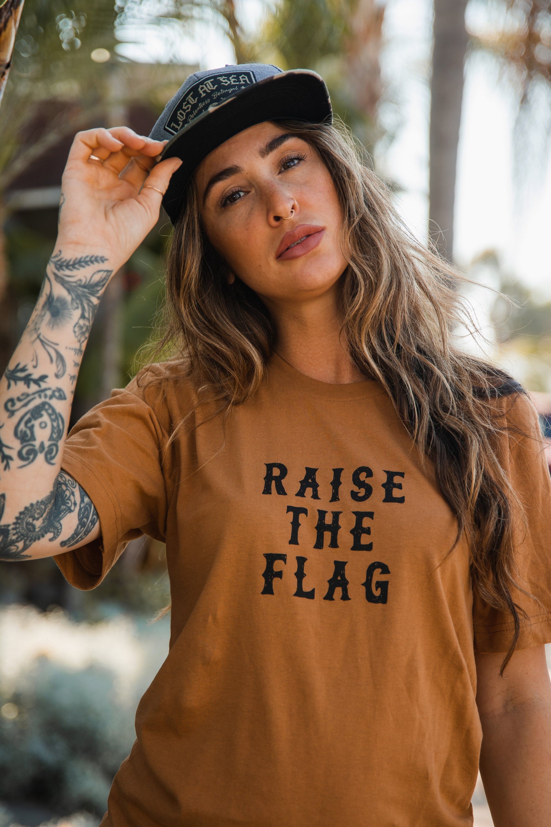 Sporting long hair and tattoos, a person wears a Relentless Betrayal Raise the Flag Tee, paired with a black cap. Outdoors against blurred greenery, they adjust their cap while making eye contact with the camera, embodying the spirit of readying to raise their own pirate flag.