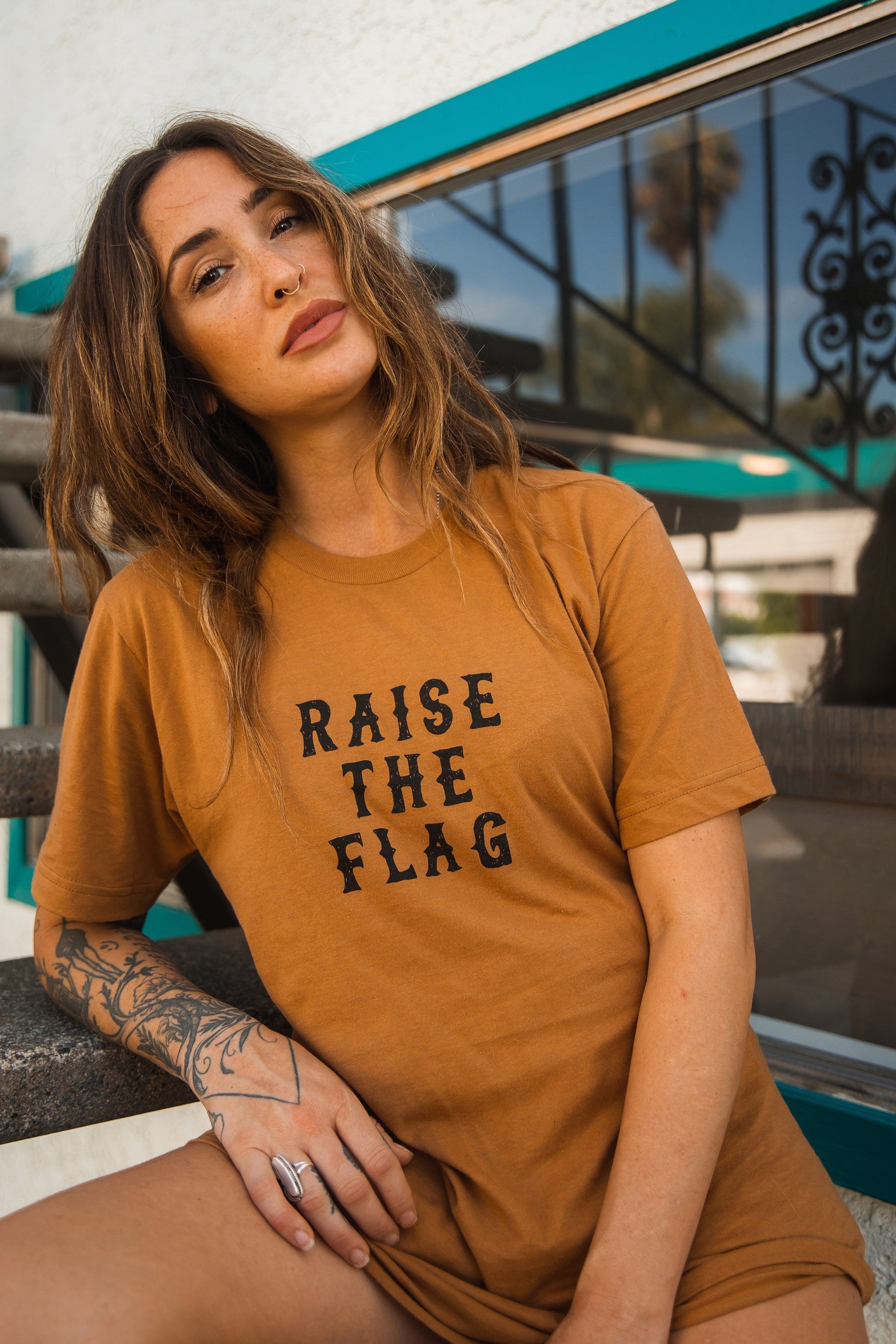 A person with long, wavy hair sits on outdoor stairs wearing the Raise the Flag Tee by Relentless Betrayal. A white building with teal trim and palm trees reflected in the window sets a backdrop that evokes a pirate flag adventure.