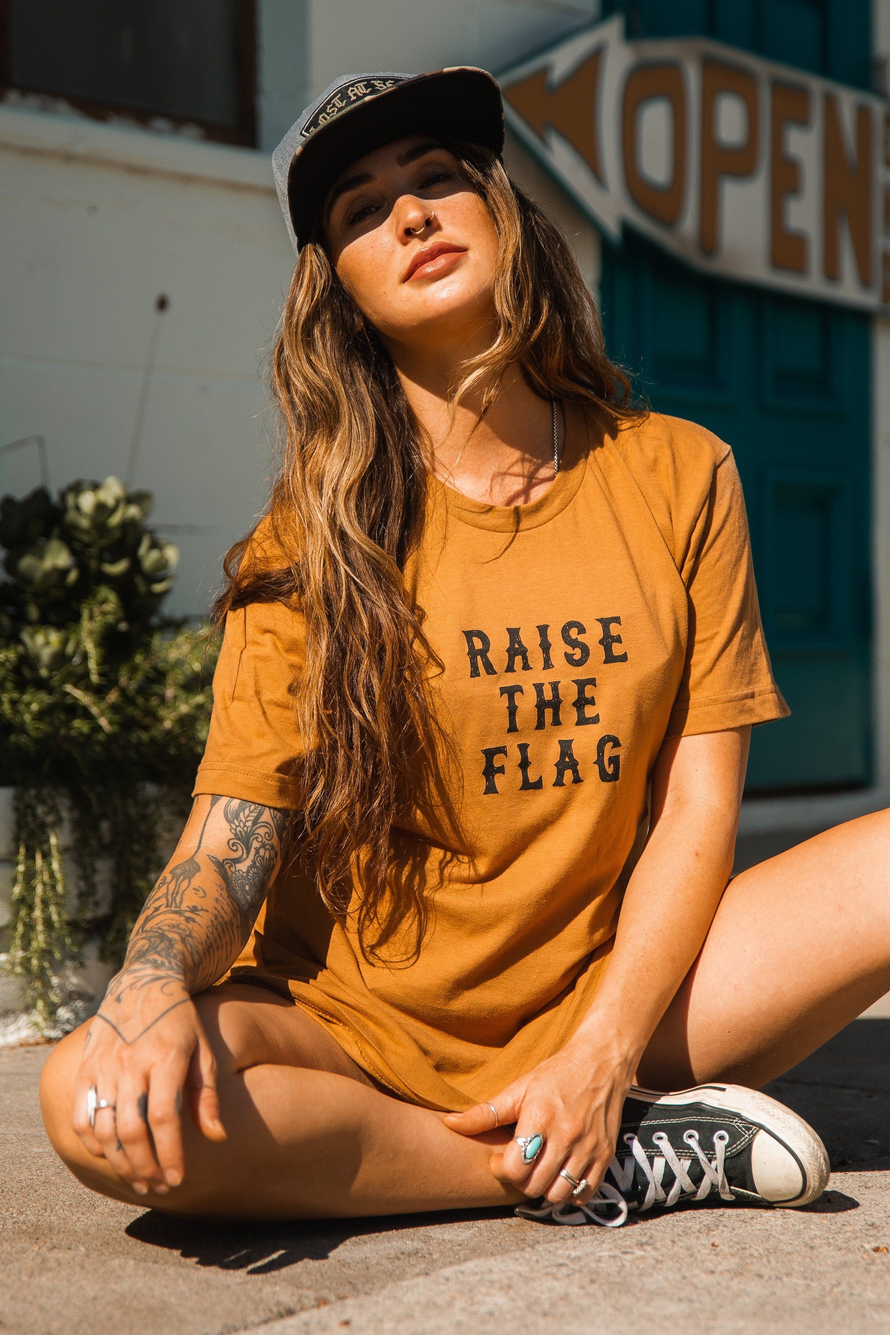 A woman sits outdoors wearing a Relentless Betrayal Raise the Flag Tee in brown jersey cotton and a cap. Her long hair falls over tattooed arms, with sneakers visible near a vintage Open sign, suggesting pirate flag tales and daring adventures.