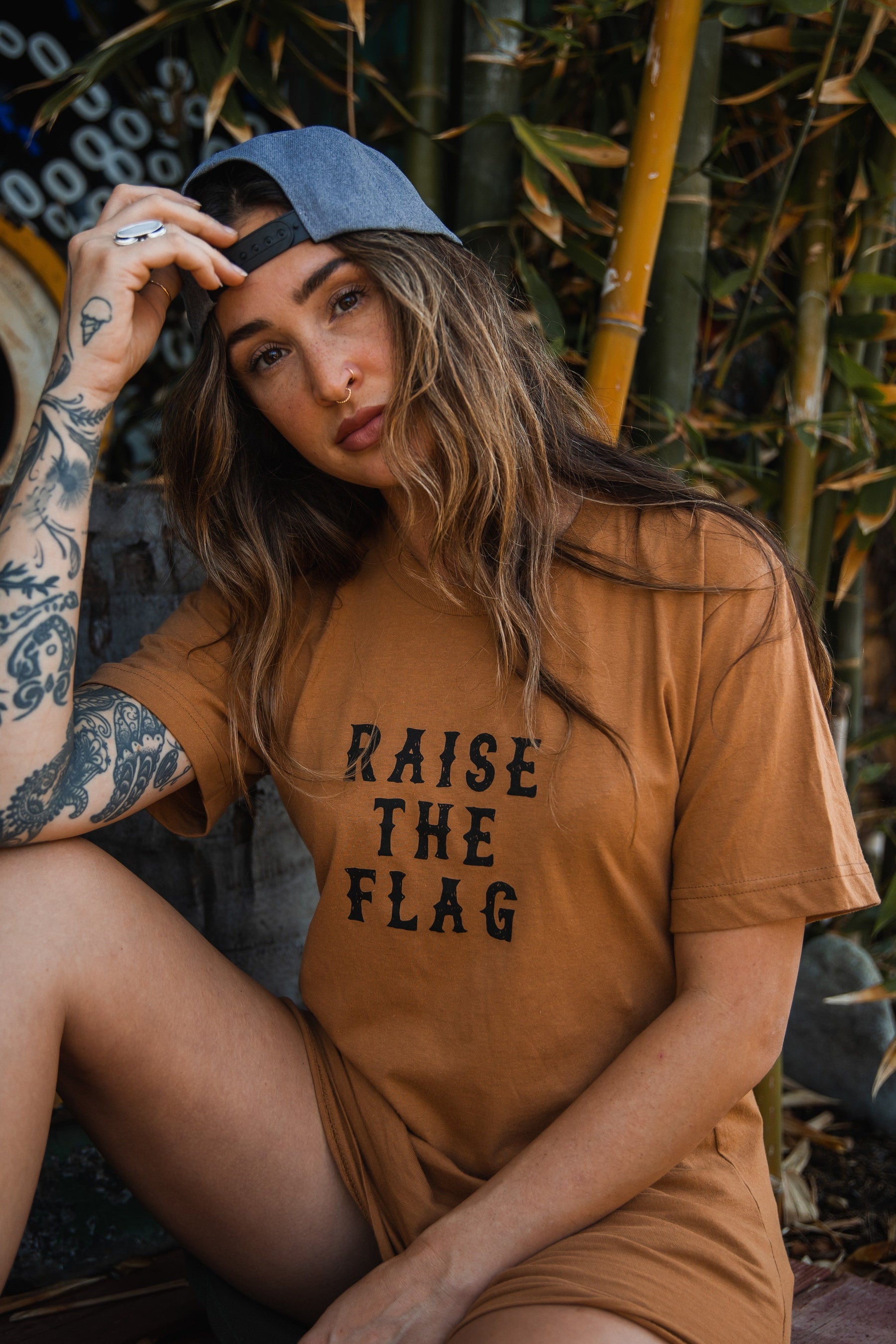 A long-haired individual relaxes outdoors with tattoos on their arm, wearing a Relentless Betrayal Raise the Flag Tee and a backward cap. Bamboo plants sway like a pirate flag in the breeze as their hand rests on the cap.