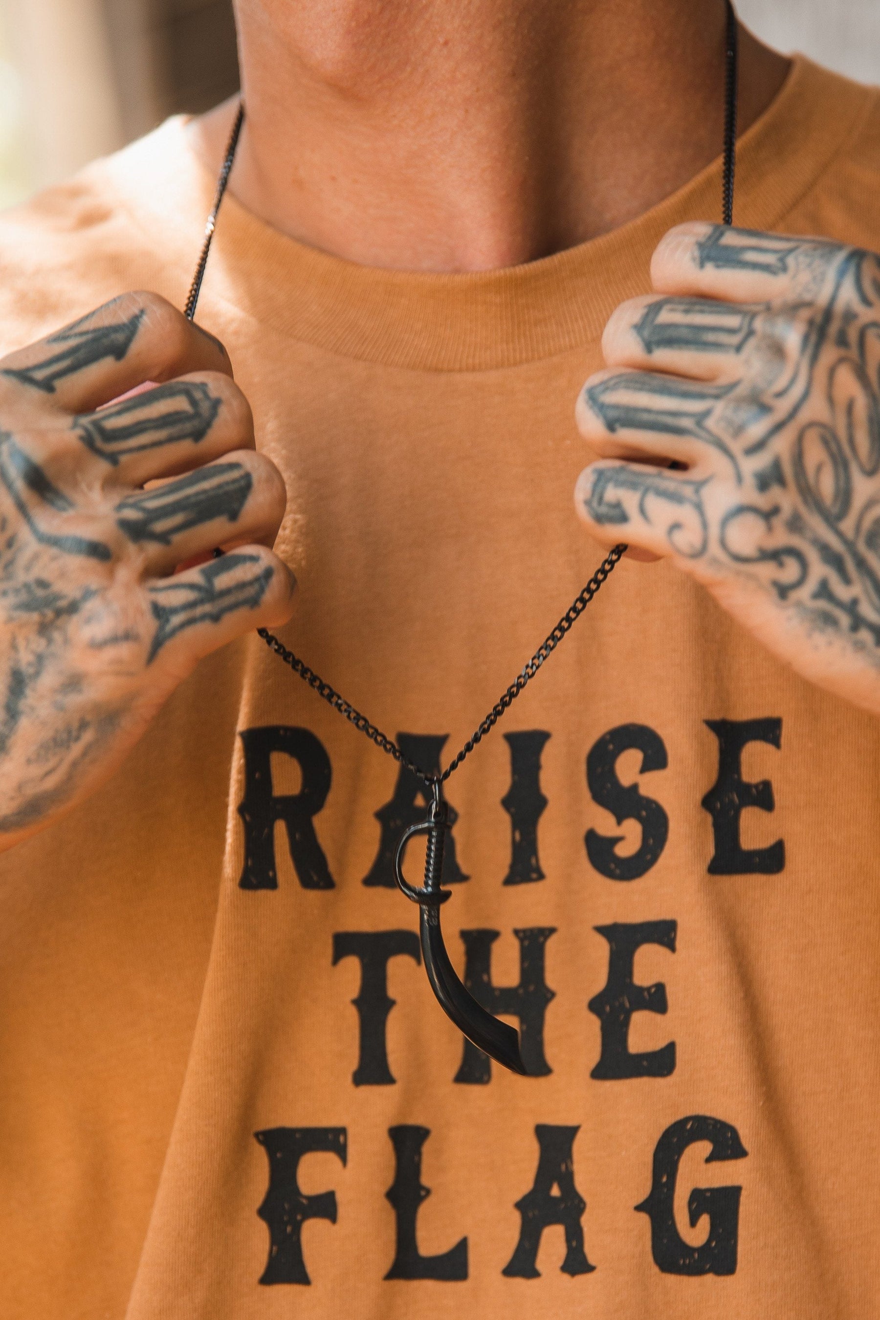 A person in a tan RAISE THE FLAG T-shirt showcases Relentless Betrayals Broadsword Necklace. Their tattooed arms contrast with the black handcrafted pendant and stainless-steel chain.