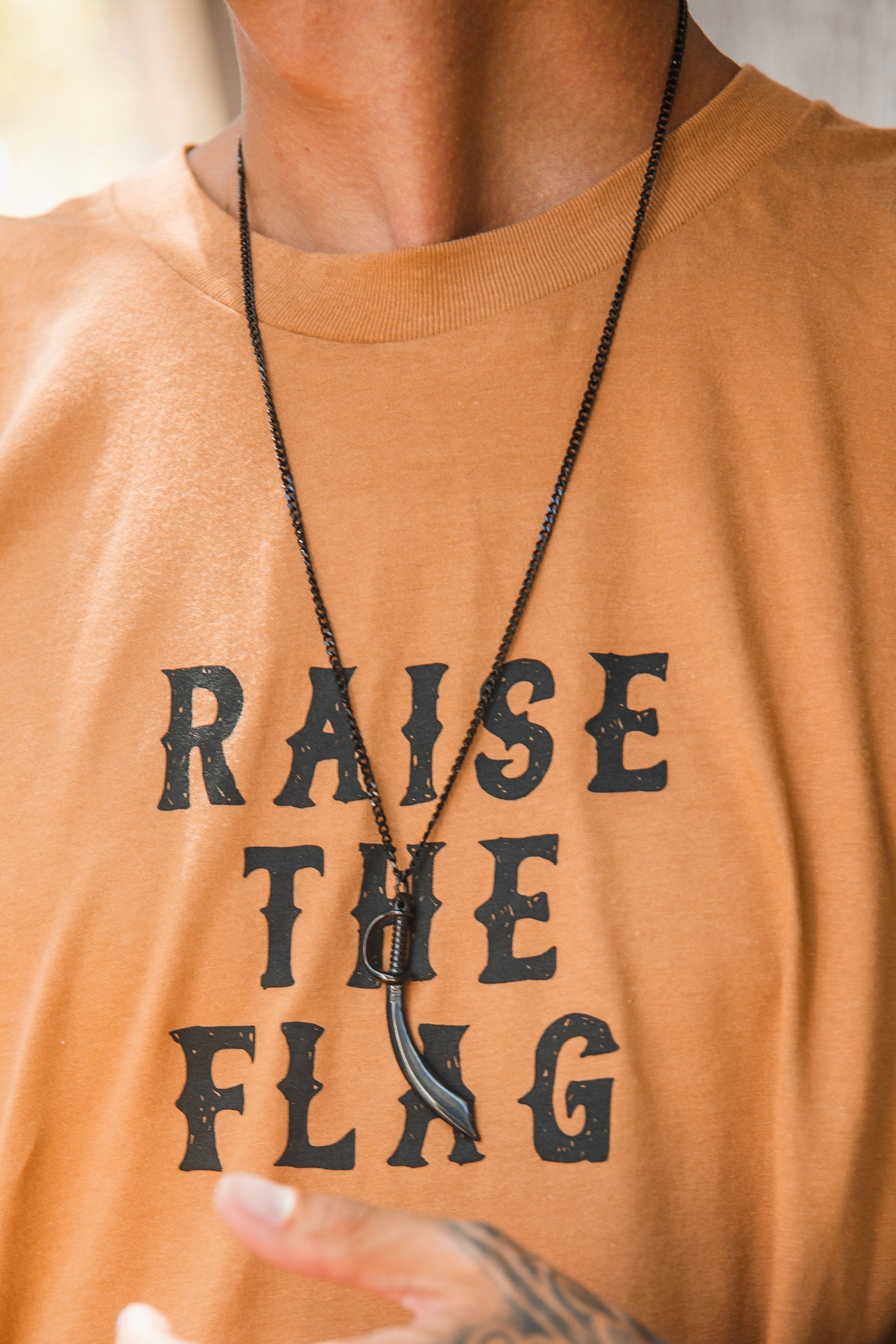 A person in a tan shirt with RAISE THE FLAG in black wears a Relentless Betrayal Broadsword Necklace on a stainless-steel chain, showcasing a hand tattoo.