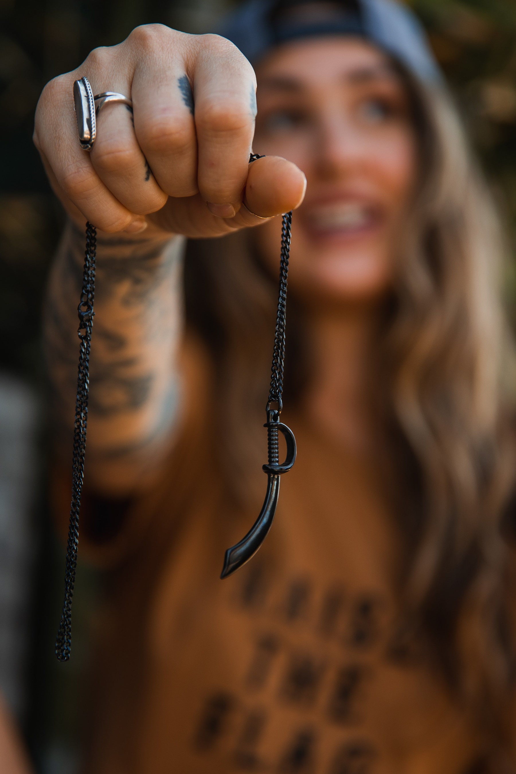 A tattooed person in casual wear smiles and holds out the Relentless Betrayals Broadsword Necklace. The handcrafted pendant with a stainless-steel chain is in focus, while the wearer is blurry in the background, wearing a cap.