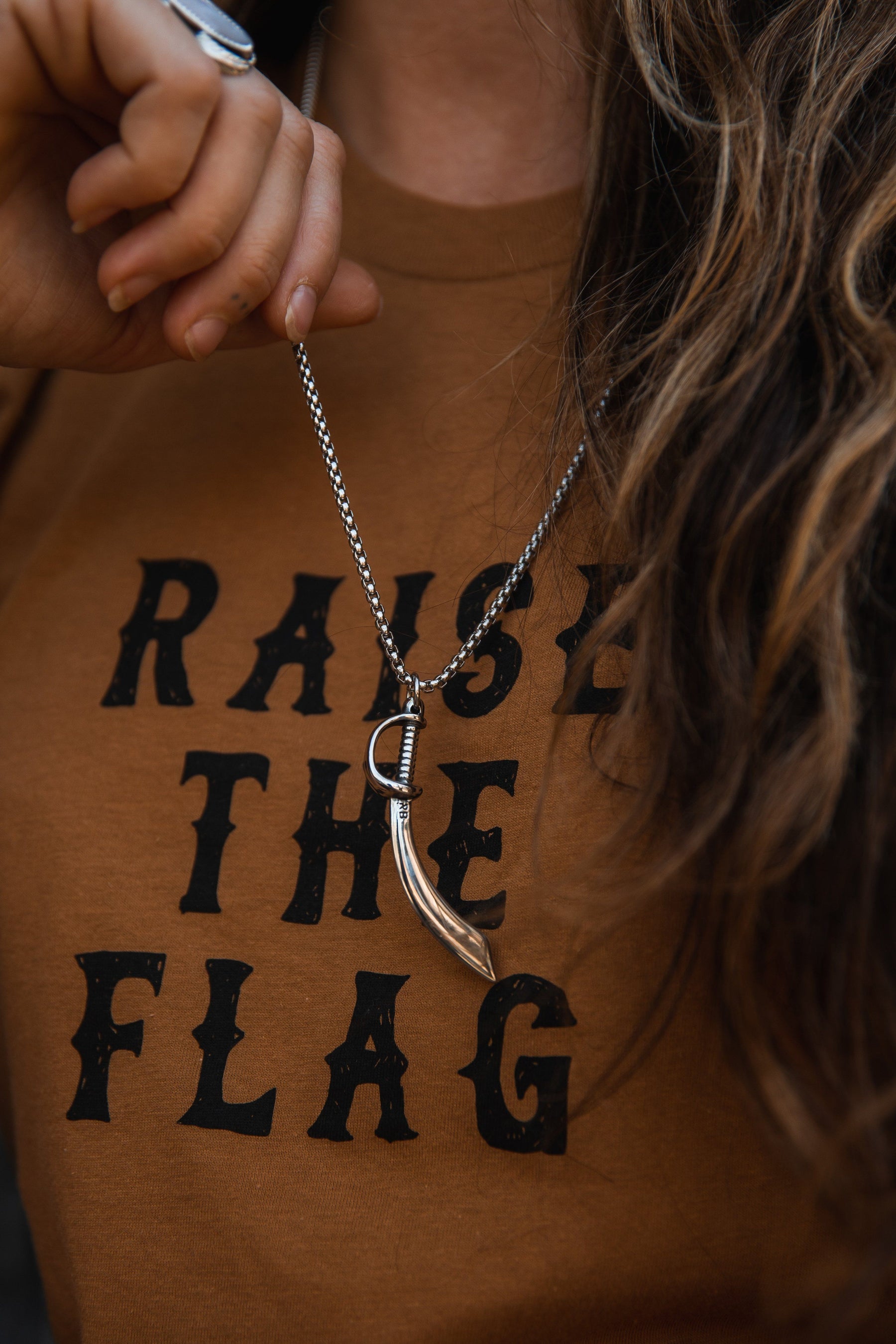 A person wearing a brown shirt with RAISE THE FLAG holds a silver Relentless Betrayal Broadsword Necklace. Their hair is wavy.