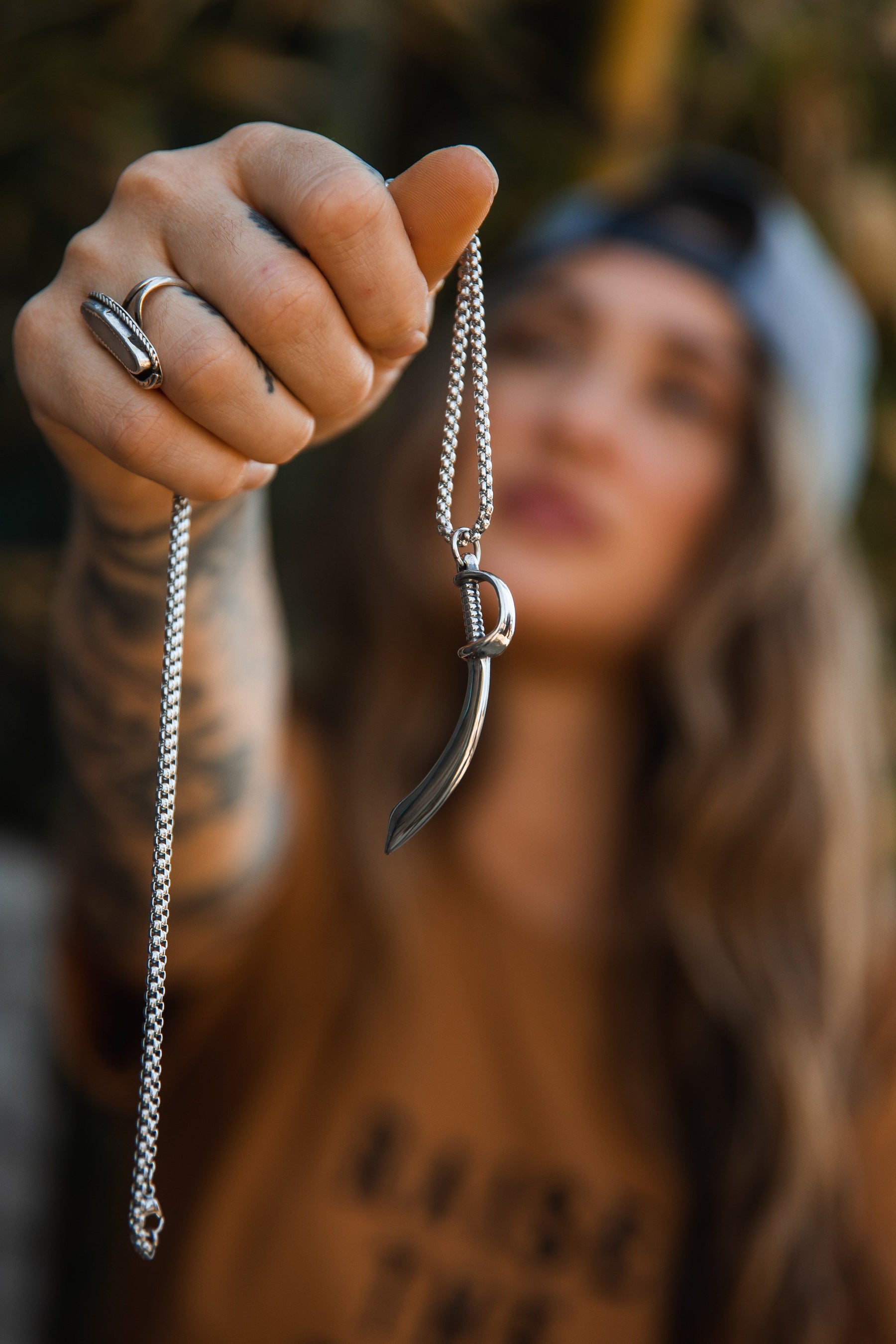 A person with long hair and a cap showcases the Relentless Betrayal Broadsword Necklace, a stainless-steel chain with a broadsword pendant. The blurred background emphasizes their tattooed arm, ring, and brown shirt, highlighting this unique handcrafted accessory.