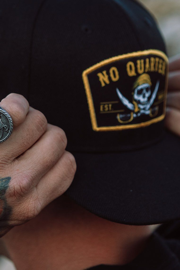 A person adjusts the Relentless Betrayal No Quarter Snapback, featuring a yellow pirate-themed patch with NO QUARTER, a skull, crossed swords, and EST. Tattooed hands and a nautical ring enhance the maritime vibe of the black flat-brim cap.