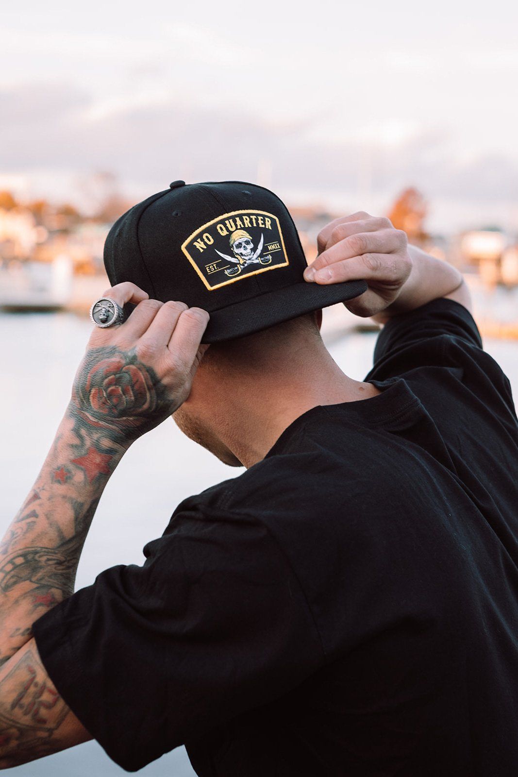 With tattooed arms, a person adjusts a Relentless Betrayal No Quarter Snapback featuring a skull and crossbones. They stand outdoors against a backdrop of blurred water and buildings.