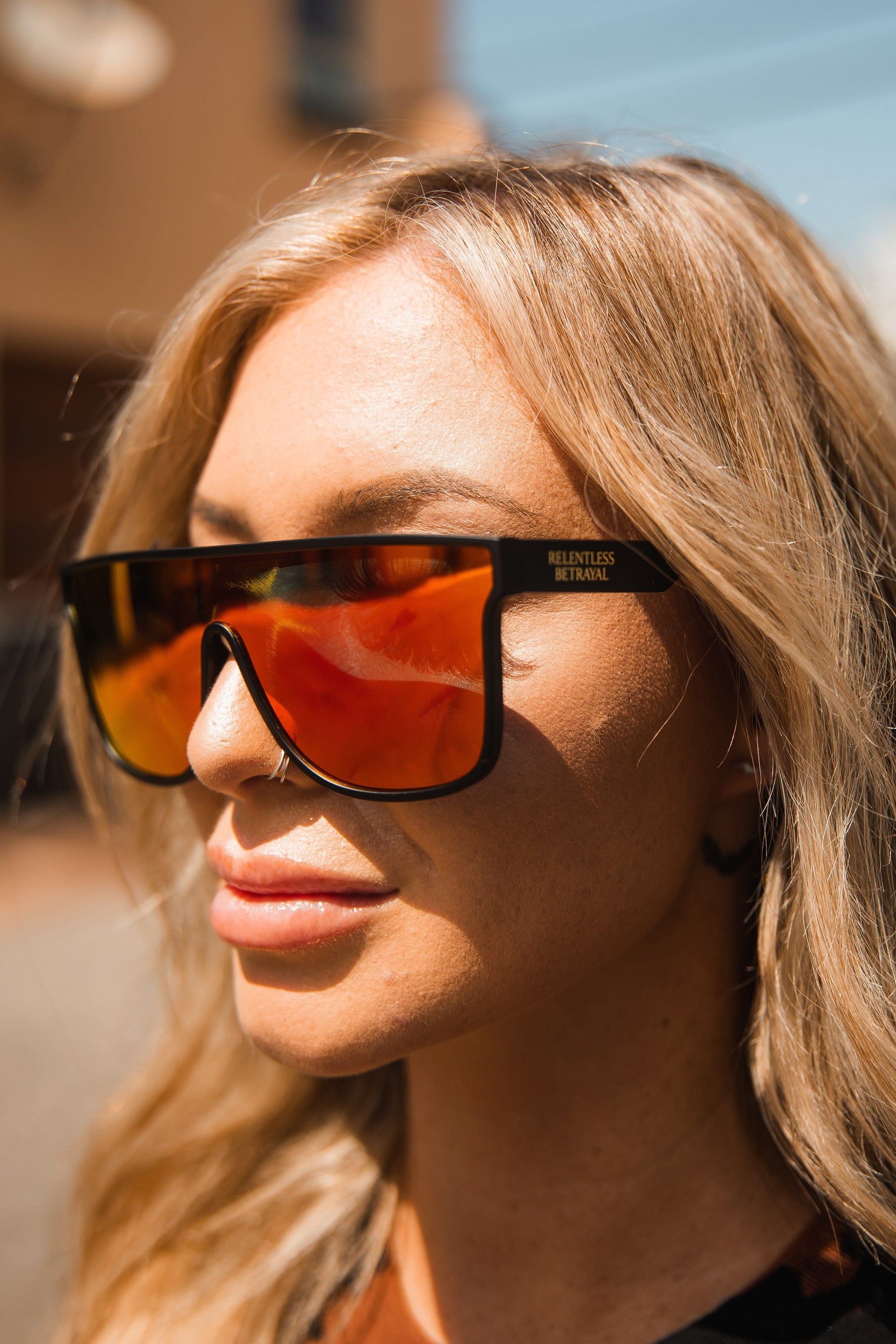 A person with long blonde hair wears RB Polarized Shades by Relentless Betrayal, looking to the side. The sun casts a warm glow on their face, highlighting the mirrored lenses. Background is blurred.