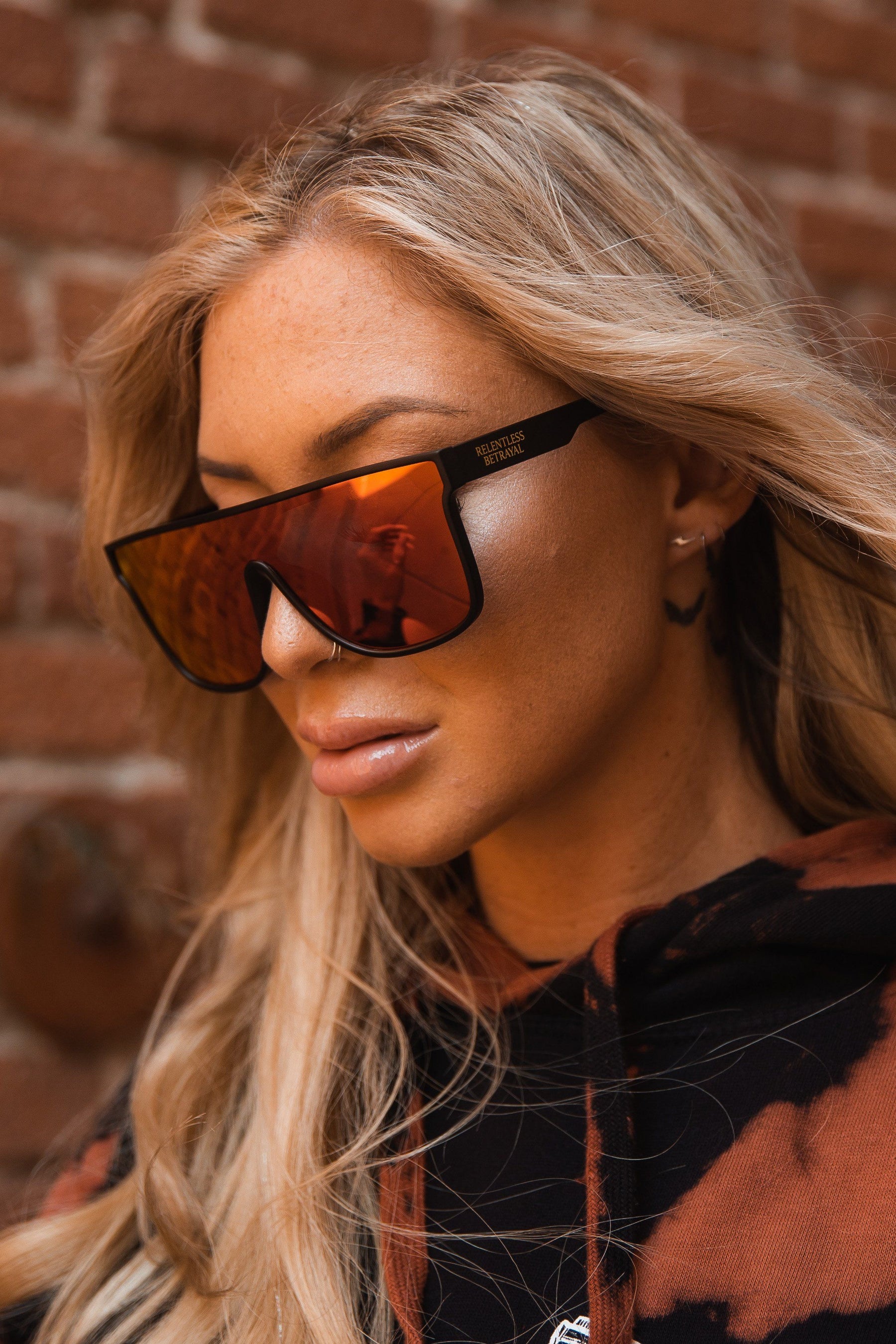 A woman with long blonde hair wears RB Polarized Shades by Relentless Betrayal, featuring large angular frames and an orange reflective tint. Shes dressed in a dark hoodie with orange patterns, standing against a rough brick wall and looking downward.