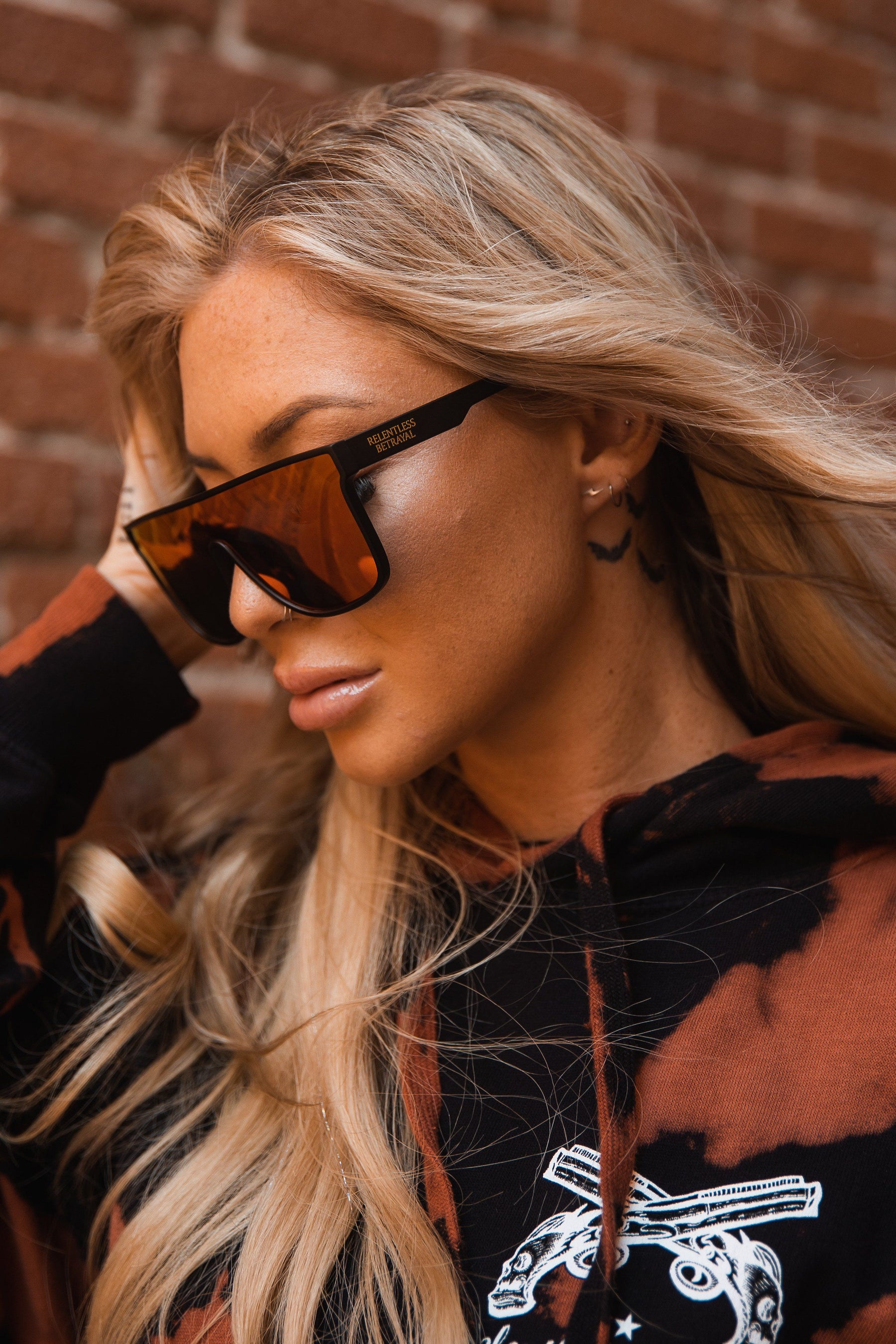 A woman with long blonde hair wearing large angular RB Polarized Shades by Relentless Betrayal and a black and brown tie-dye hoodie stands in front of a brick wall, touching her hair while looking downwards.