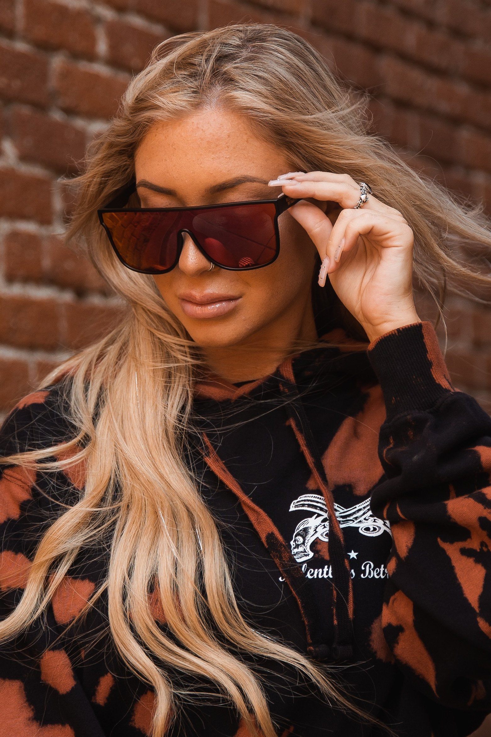 A woman with long blonde hair wears Relentless Betrayals RB Polarized Shades and a black-and-orange tie-dye hoodie. She adjusts her stylish eyewear, standing against a brick wall and gazing forward.