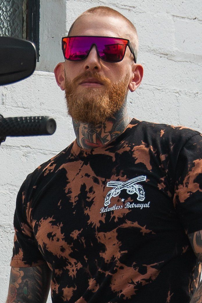 A tattooed person with a beard wears RB Polarized Shades and a black and orange Relentless Betrayal tie-dye shirt featuring crossed guns, standing out strikingly against a white brick wall.