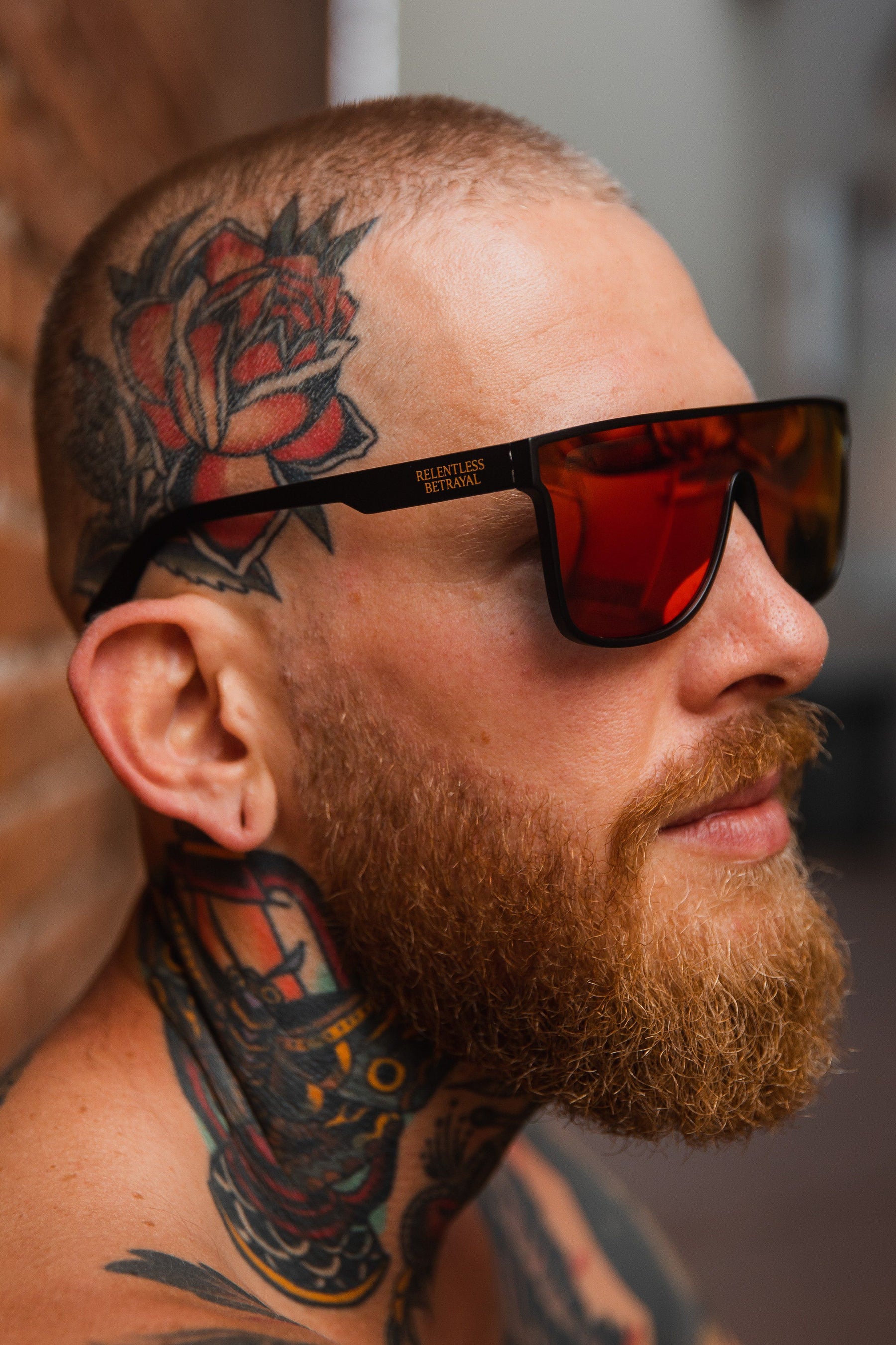 A tattooed man poses against a brick wall wearing dark RB Polarized Shades by Relentless Betrayal. He sports a rose tattoo on his head and colorful designs on his neck.
