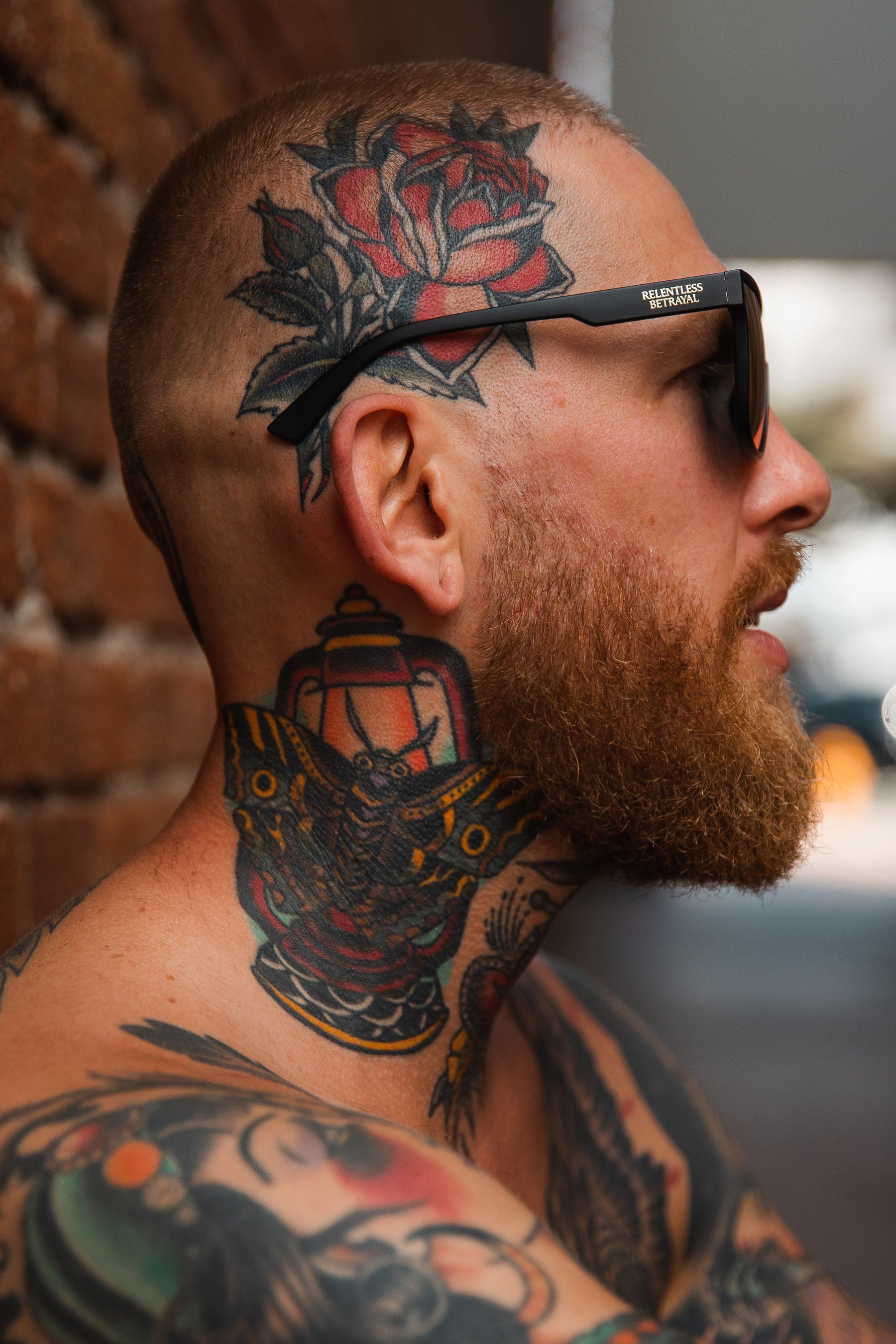 A man with a shaved head and beard leans against a brick wall while wearing RB Polarized Shades by Relentless Betrayal. His tattoos include a rose on his head, a winged lantern on his neck, and additional designs on his shoulder and arm.