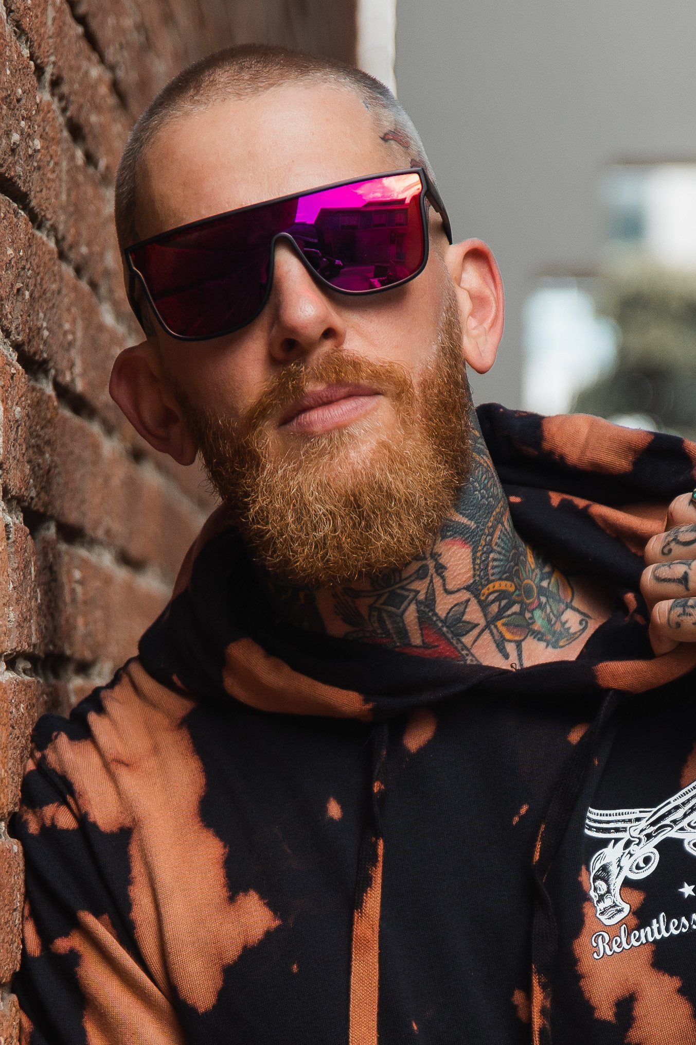 A person with a red beard and RB Polarized Shades by Relentless Betrayal leans against a brick wall. Theyre wearing a black and orange tie-dye hoodie with intricate graphic designs, showcasing tattoos on their neck and hands.