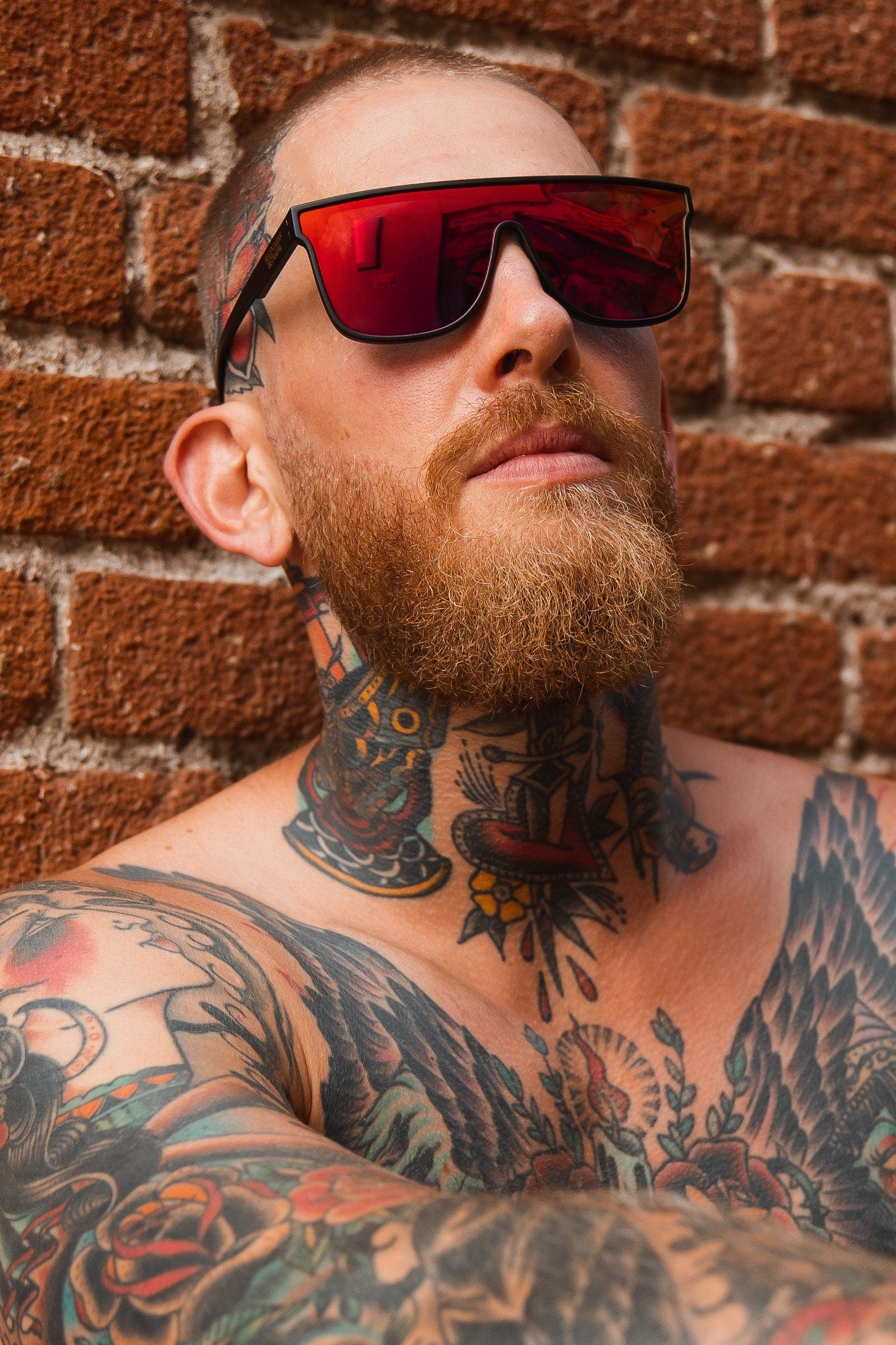 A bearded man with colorful chest and neck tattoos leans against a brick wall, wearing Relentless Betrayals RB Polarized Shades, boasting UV protection. He gazes upward with confidence.