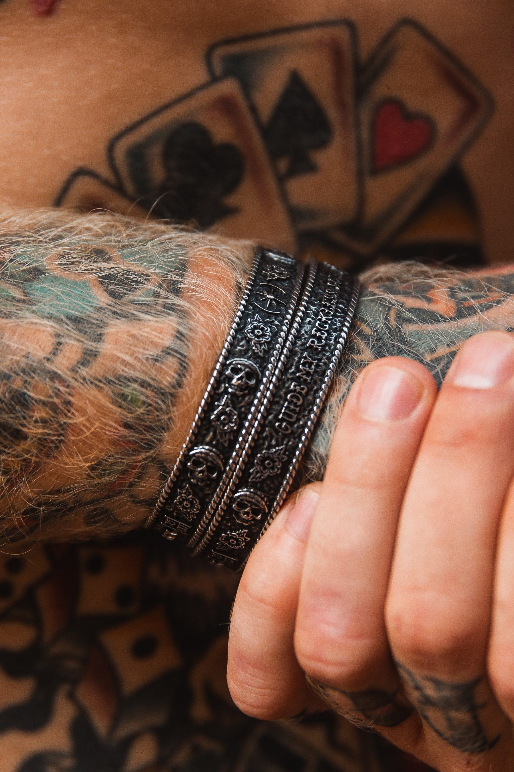 A close-up of a tattooed arm showcases the elegance of Relentless Betrayals Guide My Reckless Soul Bracelet, one of three detailed, handcrafted pieces. In the background, playing cards decorate the chest as a partially visible hand is slightly clenched at the bottom right.