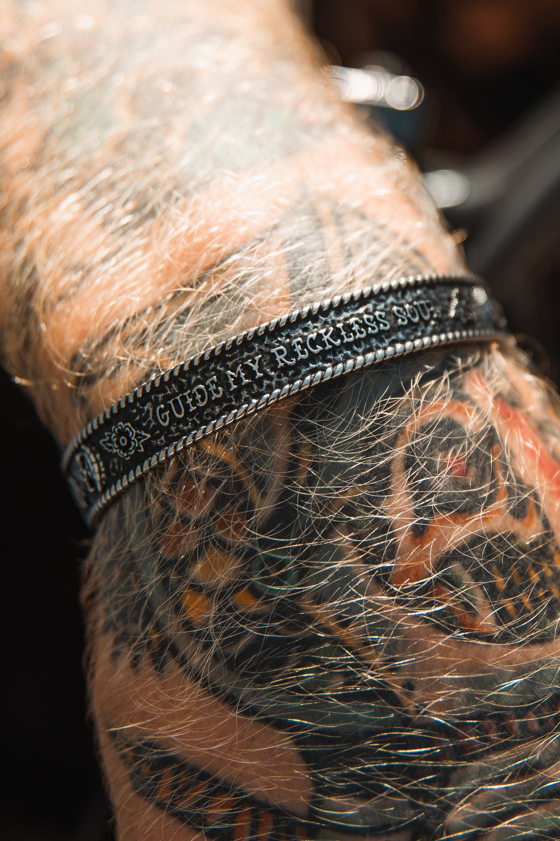 A tattooed arm wears the Guide My Reckless Soul Bracelet by Relentless Betrayal, featuring engraved text and decorative elements with an adjustable fit.