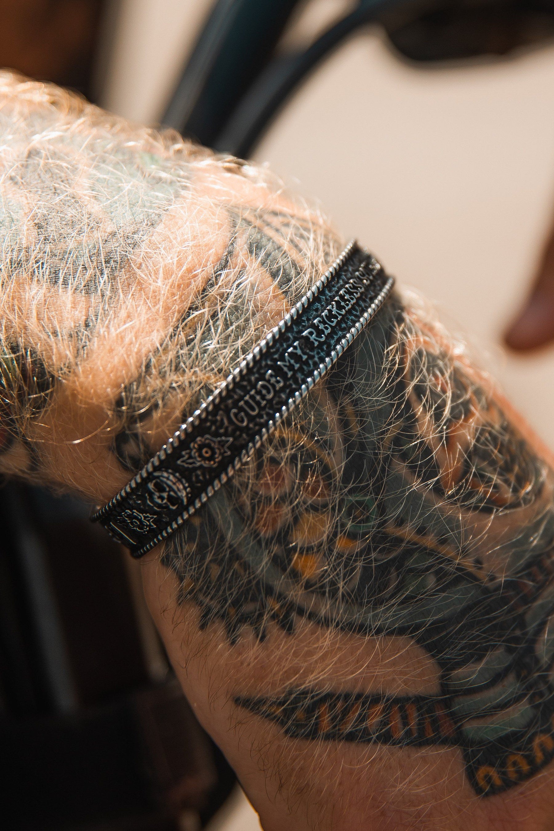 Close-up of a forearm with intricate bird head tattoos, adorned with the Guide My Reckless Soul Bracelet by Relentless Betrayal. This handcrafted black bracelet with detailed patterns stands out against light brown, sunlit, hairy skin, highlighting both the tattoo and bracelet artistry.