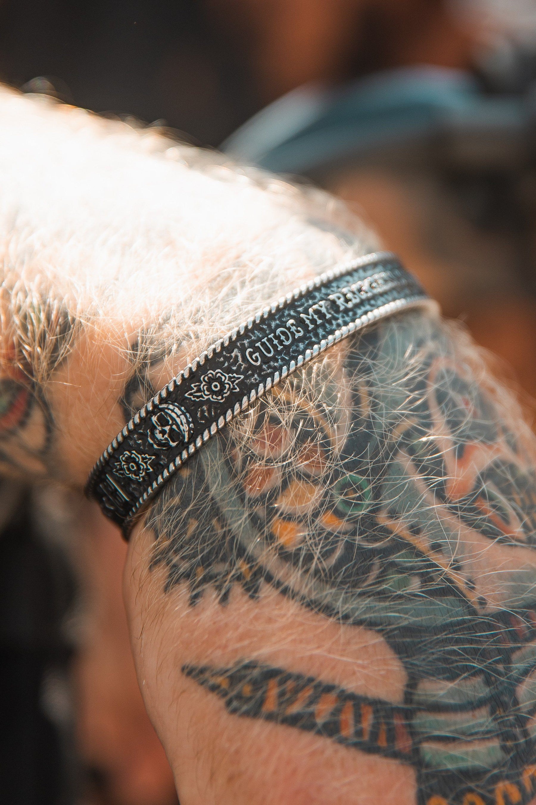A tattooed arm wears an adjustable black Guide My Reckless Soul bracelet by Relentless Betrayal. The colorful tattoos and gentle light enhance the handcrafted stainless steel details.