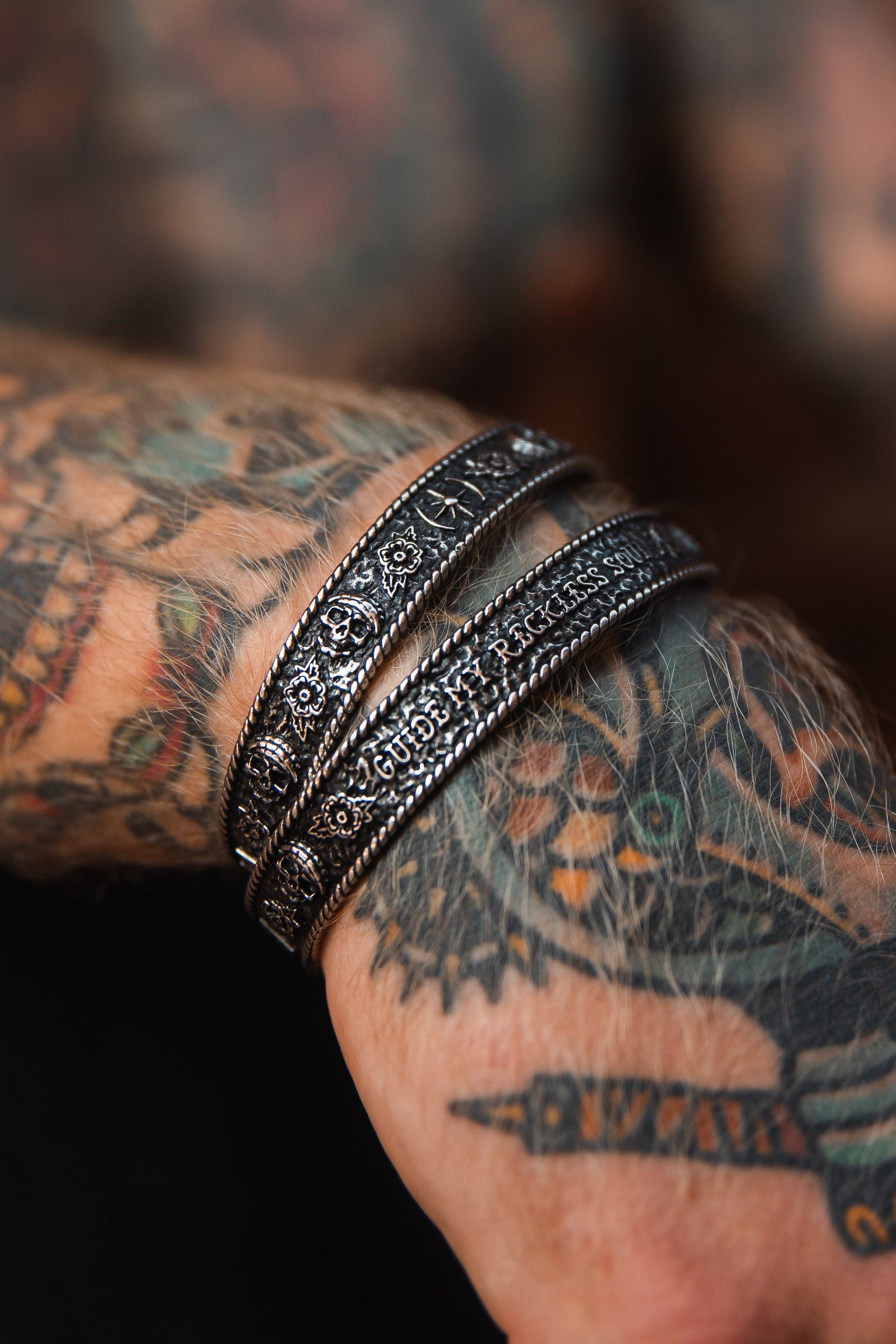A tattooed forearm showcases two ornate Guide My Reckless Soul Bracelets by Relentless Betrayal. The bracelets, crafted in engraved silver with an adjustable fit, complement the colorful and detailed tattoos with a handcrafted and edgy look.