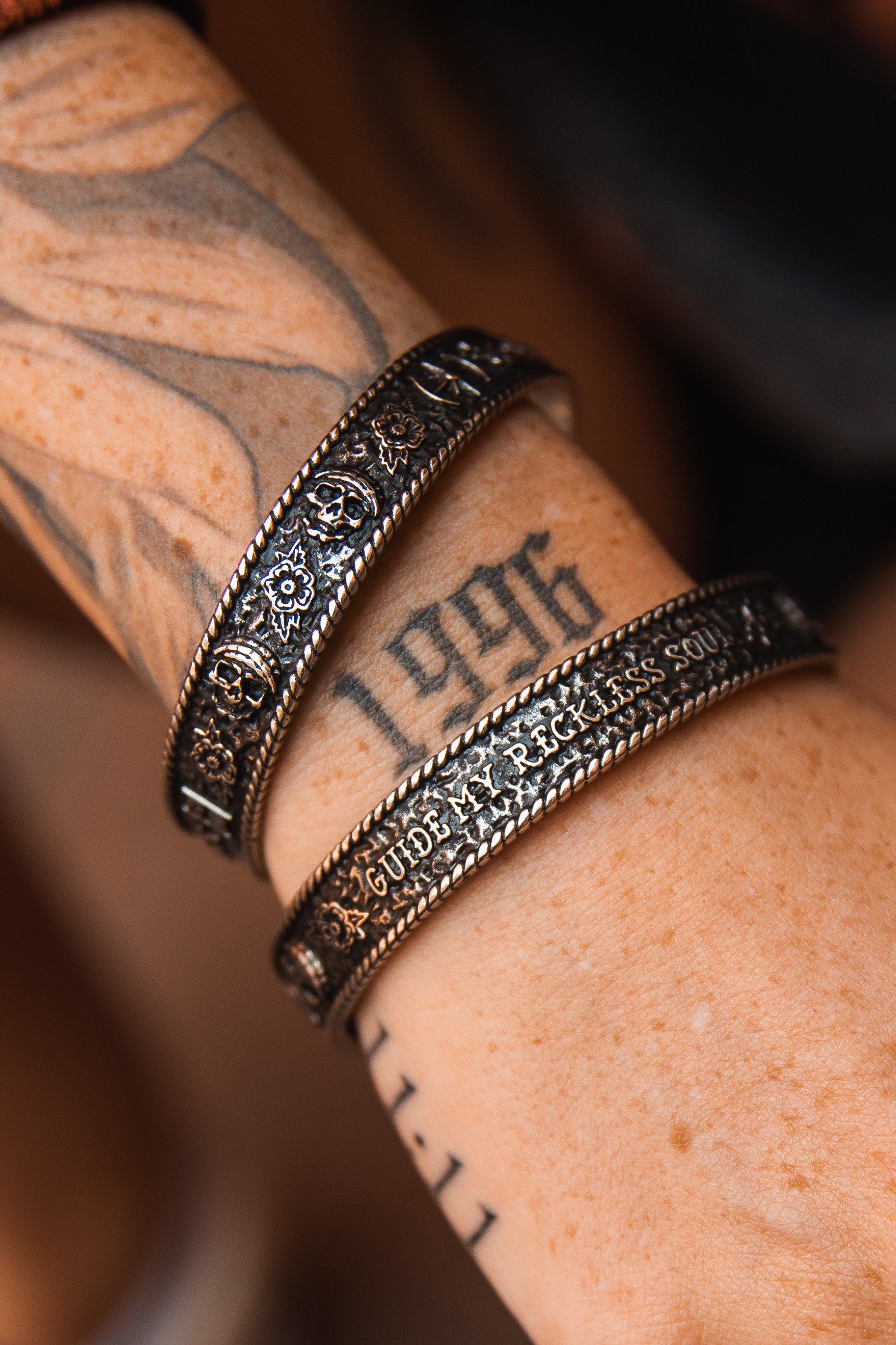 A close-up of a tattooed forearm with 1996 and GUIDE, adorned with two ornate, handcrafted metal bracelets. The Guide My Reckless Soul Bracelet by Relentless Betrayal features intricate patterns and textures, offering an adjustable fit for comfort.