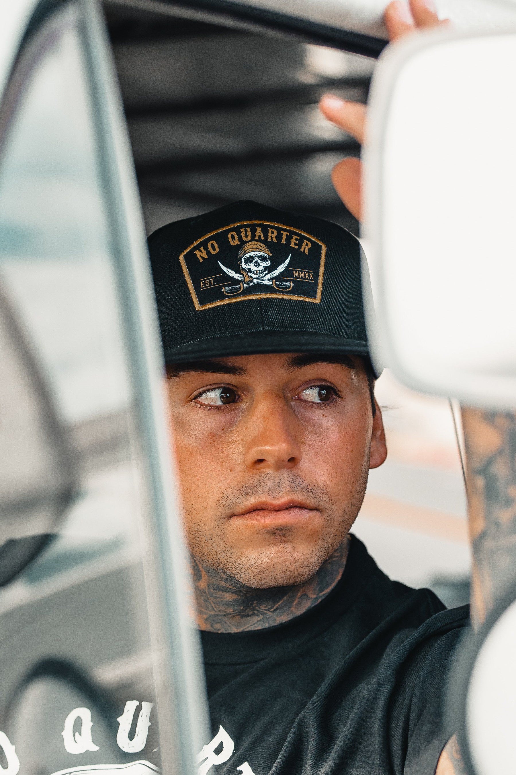 A tattooed individual wears a Relentless Betrayal black No Quarter Snapback, featuring a skull and crossbones, looking to the side while seated in a vehicle. They grip a white object, possibly part of the visor or window frame.