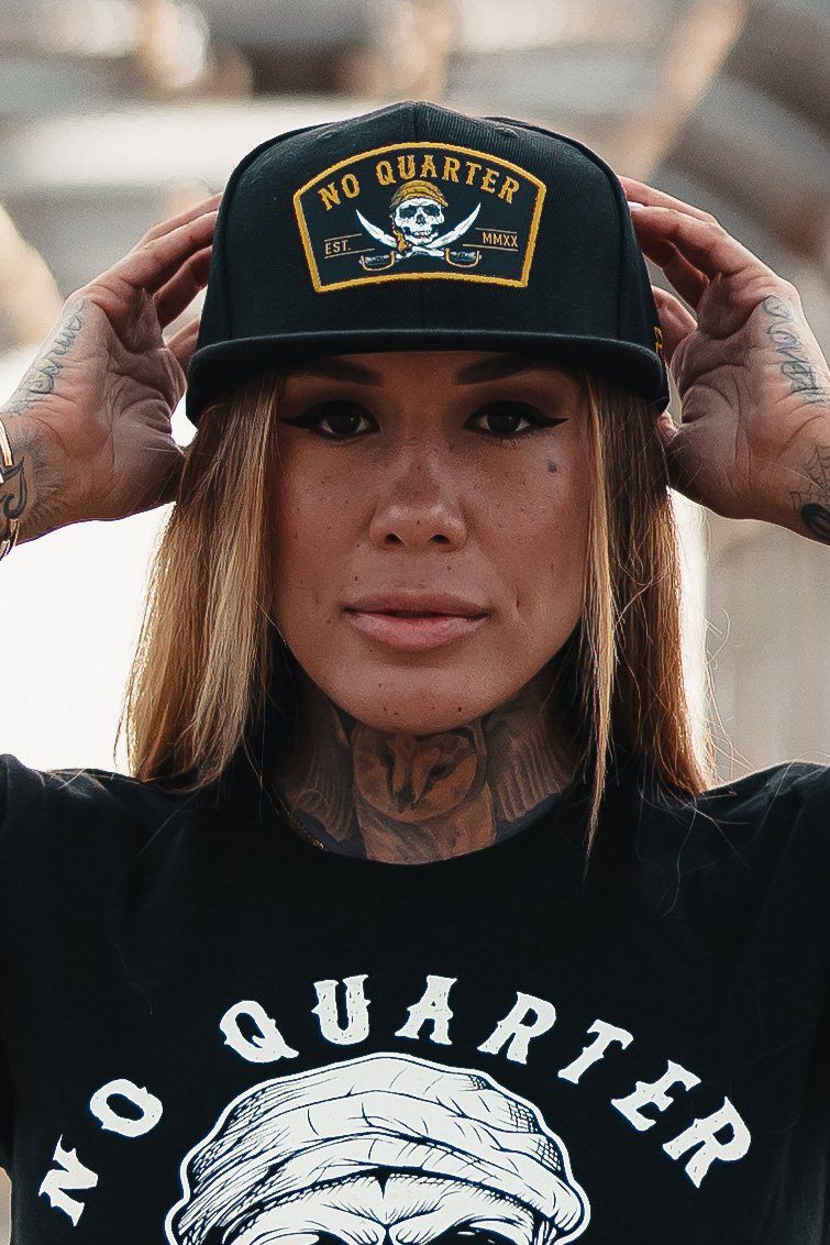 Sporting a No Quarter snapback from Relentless Betrayal, a person poses with one hand on the brim. The hat features a skull and crossbones design, complementing their tats on hands and neck, as they maintain a neutral expression.