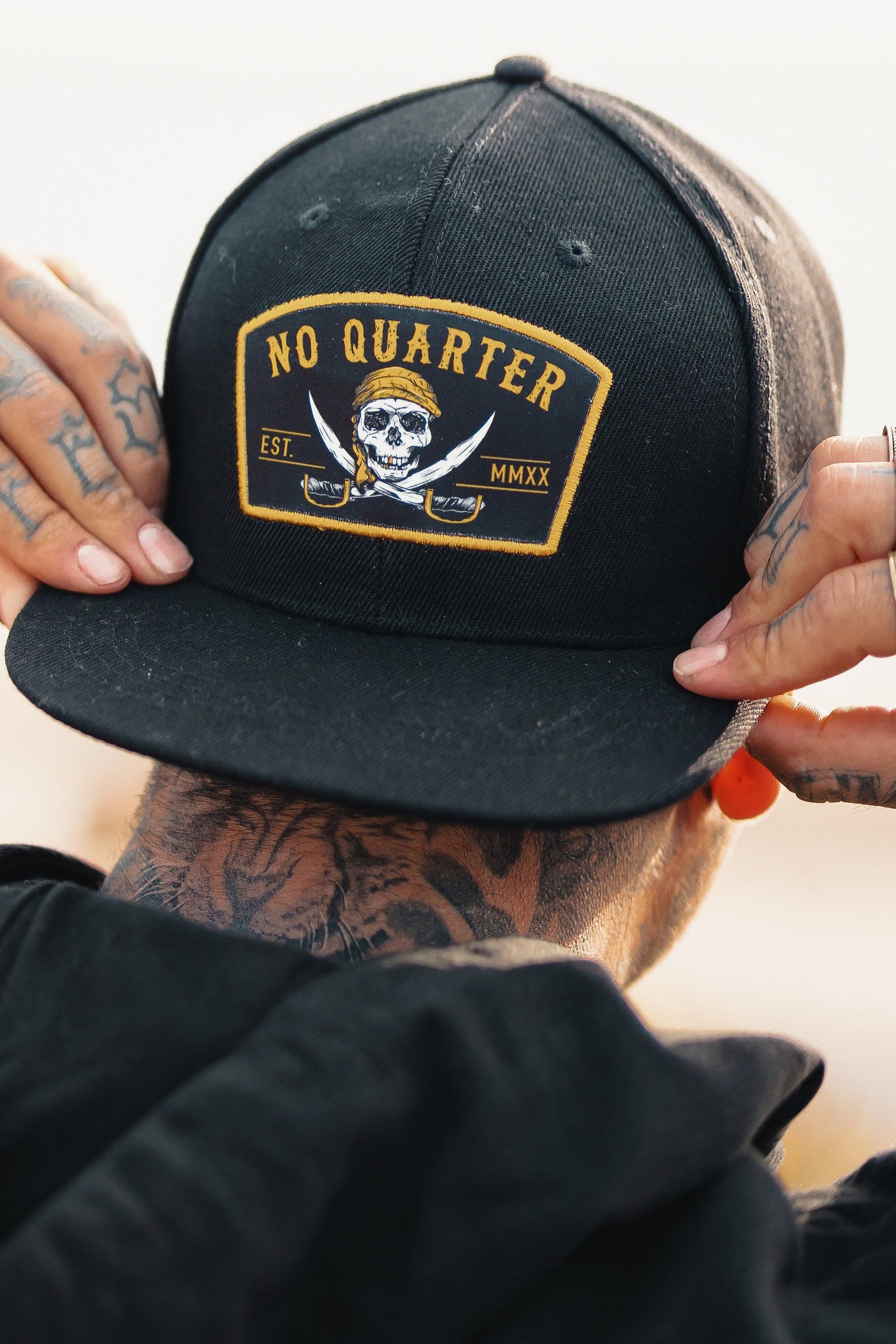 A person adjusts a Relentless Betrayal No Quarter Snapback featuring a skull and crossed swords with Est. MMXX. Tattoos adorn their neck and hands, complemented by an orange plug earring.
