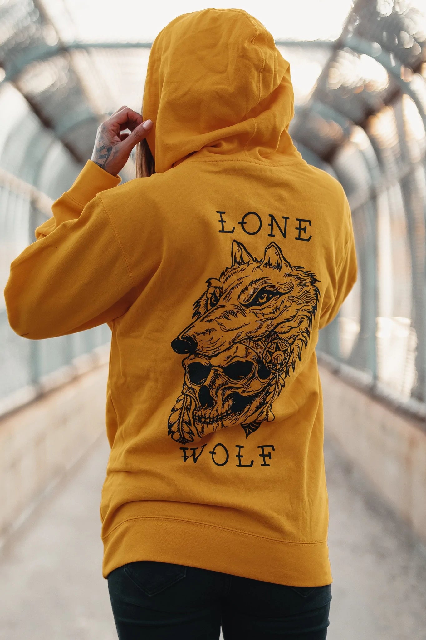A person in a Lone Wolf GOLD Hoodie by Relentless Betrayal, featuring a wolf illustration and text, stands on a pedestrian bridge with metal railings and a covered structure. The hood is up as they adjust it with one hand.