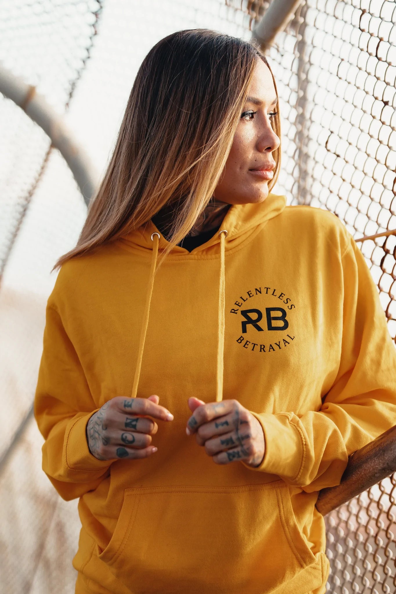 A solitary figure in a Lone Wolf GOLD Hoodie by Relentless Betrayal stands by a chain-link fence, their long hair and tattooed fingers complementing their thoughtful expression, embodying unisex style as they gaze into the distance.