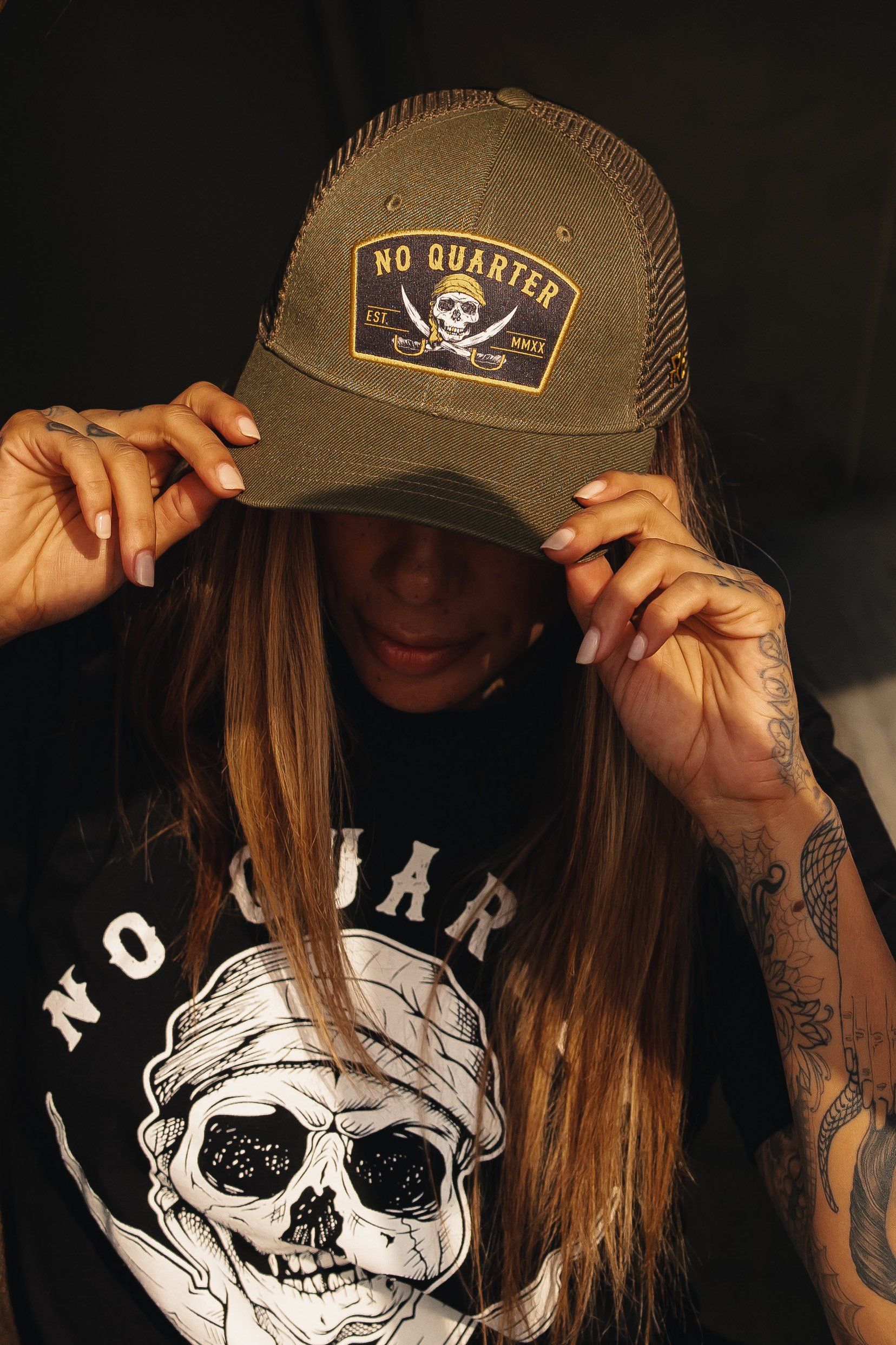 A person in a brown No Quarter Trucker Snapback by Relentless Betrayal adjusts the brim while wearing a black shirt with a similar skull graphic. Tattoos on their arms enhance their edgy appearance.