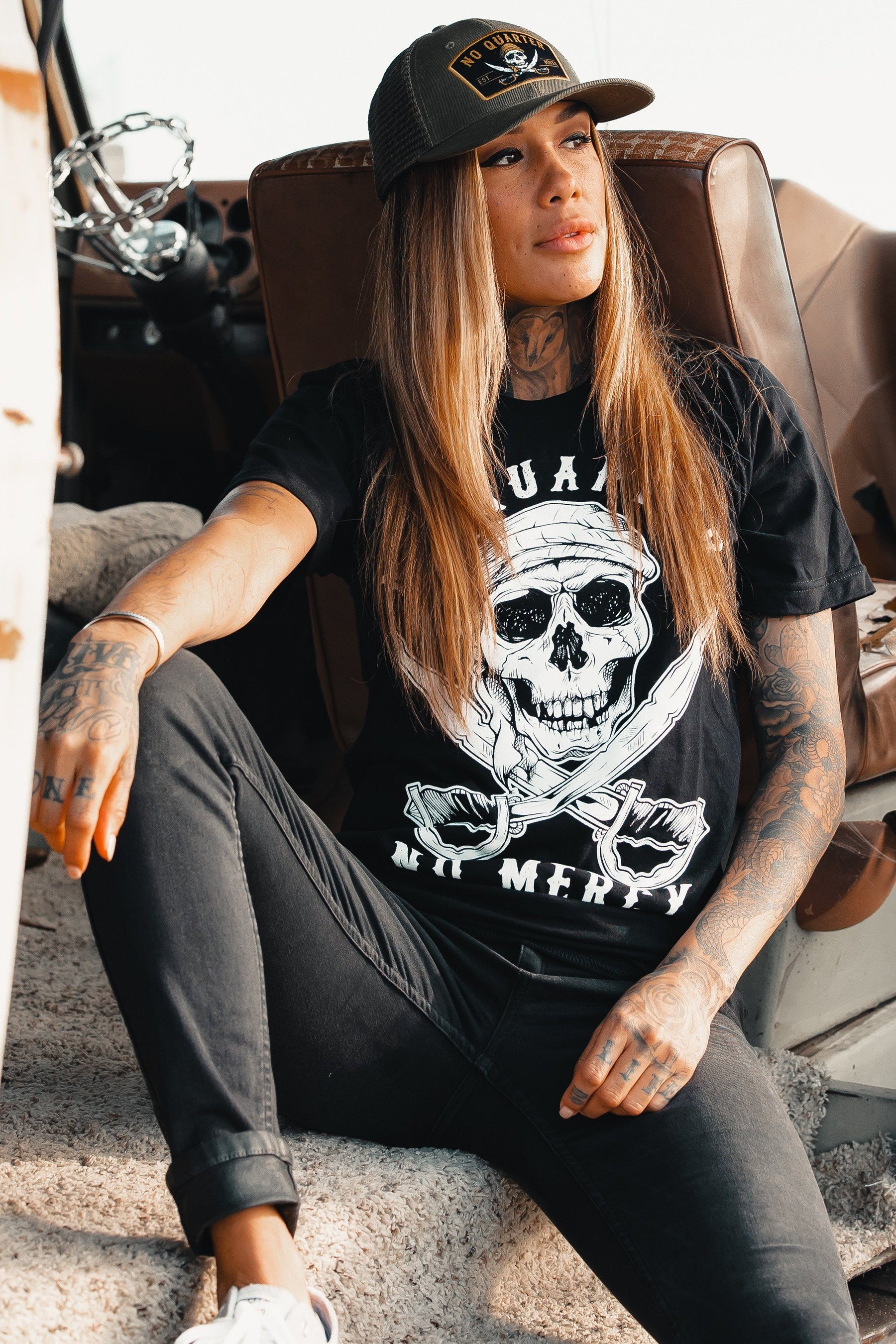 A woman sits casually with a relaxed posture in a vehicle seat, wearing the No Quarter Trucker Snapback by Relentless Betrayal and a graphic t-shirt with a skull design. Her long hair drapes over her shoulders, showing tattoos on her arms in a laid-back outdoor setting.