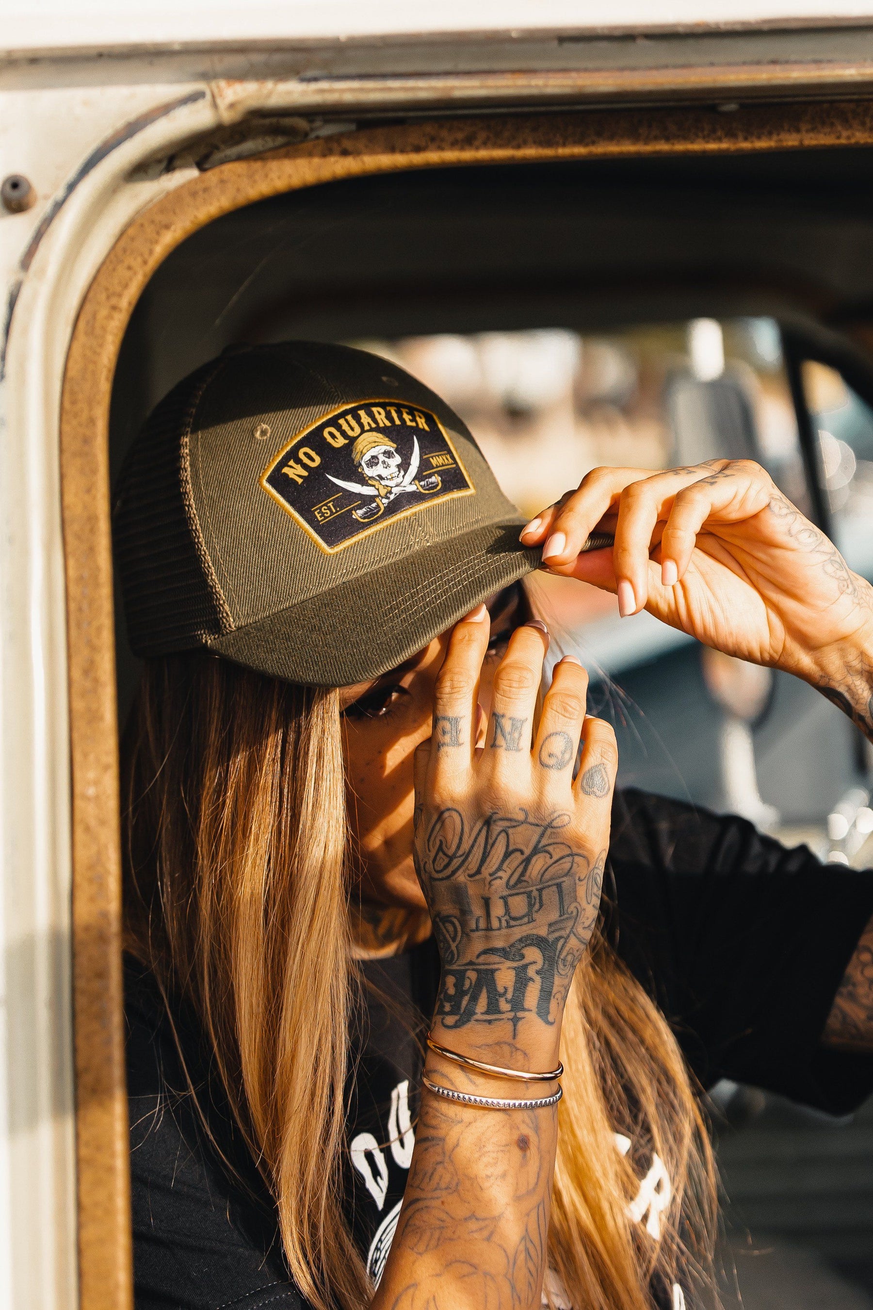 A person with long hair adjusts their Relentless Betrayal No Quarter Trucker Snapback, featuring a skull and crossbones. Leaning out the window, their intricately tattooed hand is visible.