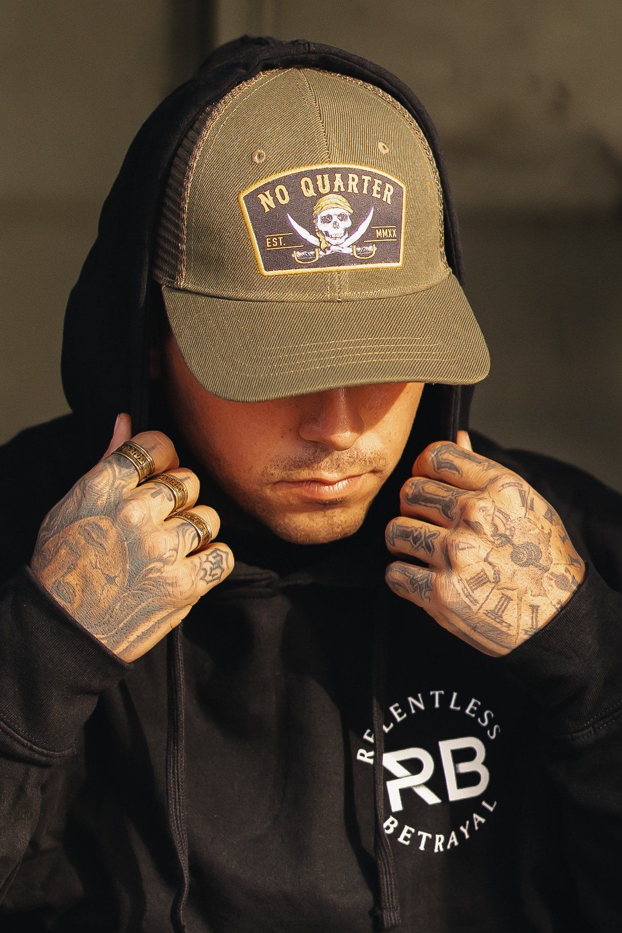 A person with tattooed hands adjusts the hood of their sweatshirt, displaying Relentless Betrayal, while wearing a green No Quarter Trucker Snapback featuring a skull logo and the brand name.