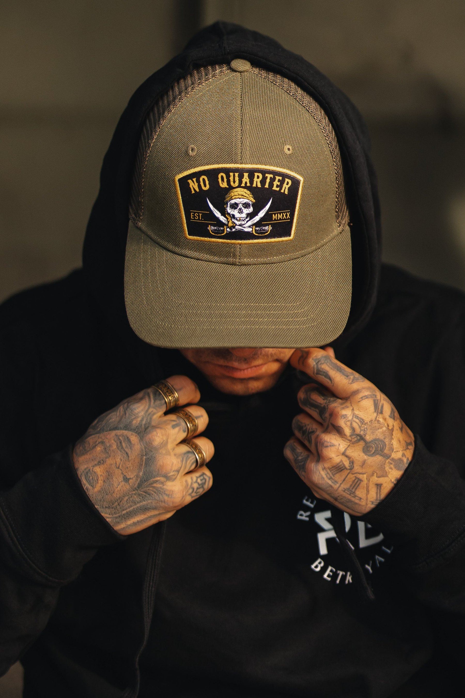 A person wearing a Relentless Betrayal No Quarter Trucker Snapback and a black hoodie, adorned with tattoos and rings, slightly tilts their head downwards.