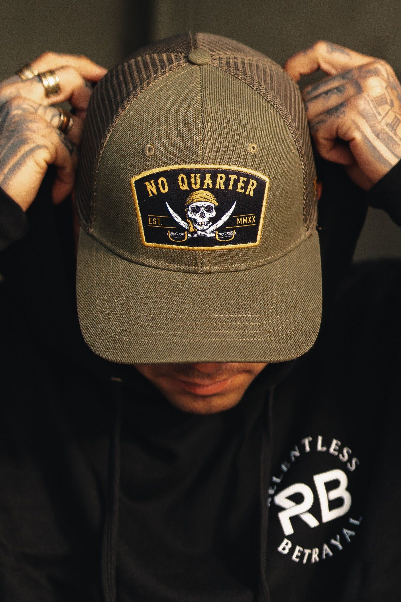 Wearing a dark green No Quarter Trucker Snapback, featuring a skull and crossbones patch, they adjust the snapback enclosure. Paired with a black Relentless Betrayal hoodie, their rings and tattoos enhance their edgy style.