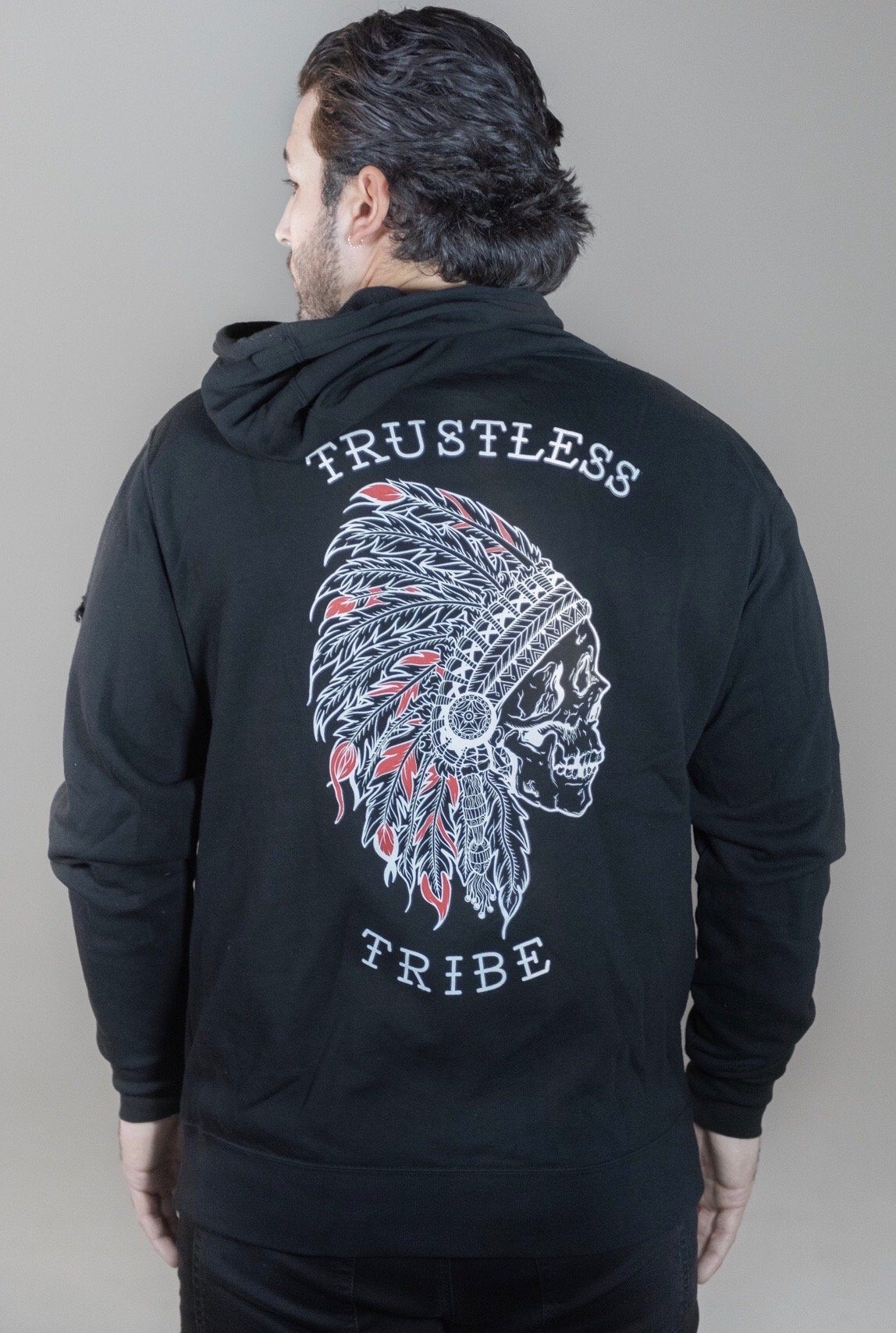 A man wearing the Relentless Betrayal Trustless Chief Hoodie RED—a unisex black hoodie featuring a skull in a feathered headdress and the words “TRUSTLESS TRIBE” printed on the back, crafted from a comfortable cotton-polyester blend.