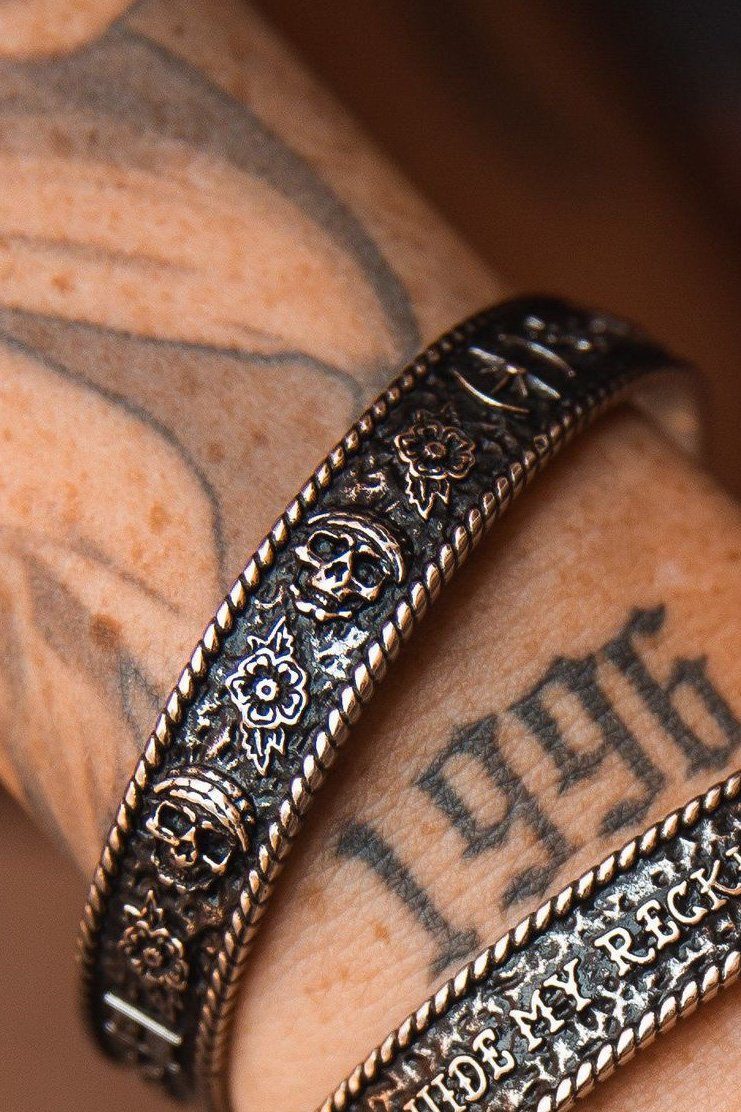 Close-up of a tattooed arm with two handcrafted metal bracelets. The Guide My Reckless Soul bracelet by Relentless Betrayal displays intricate skull and flower designs in stainless steel, while the other bracelet features engraved text. A 1996 tattoo peeks out beside the adjustable fit bracelets.