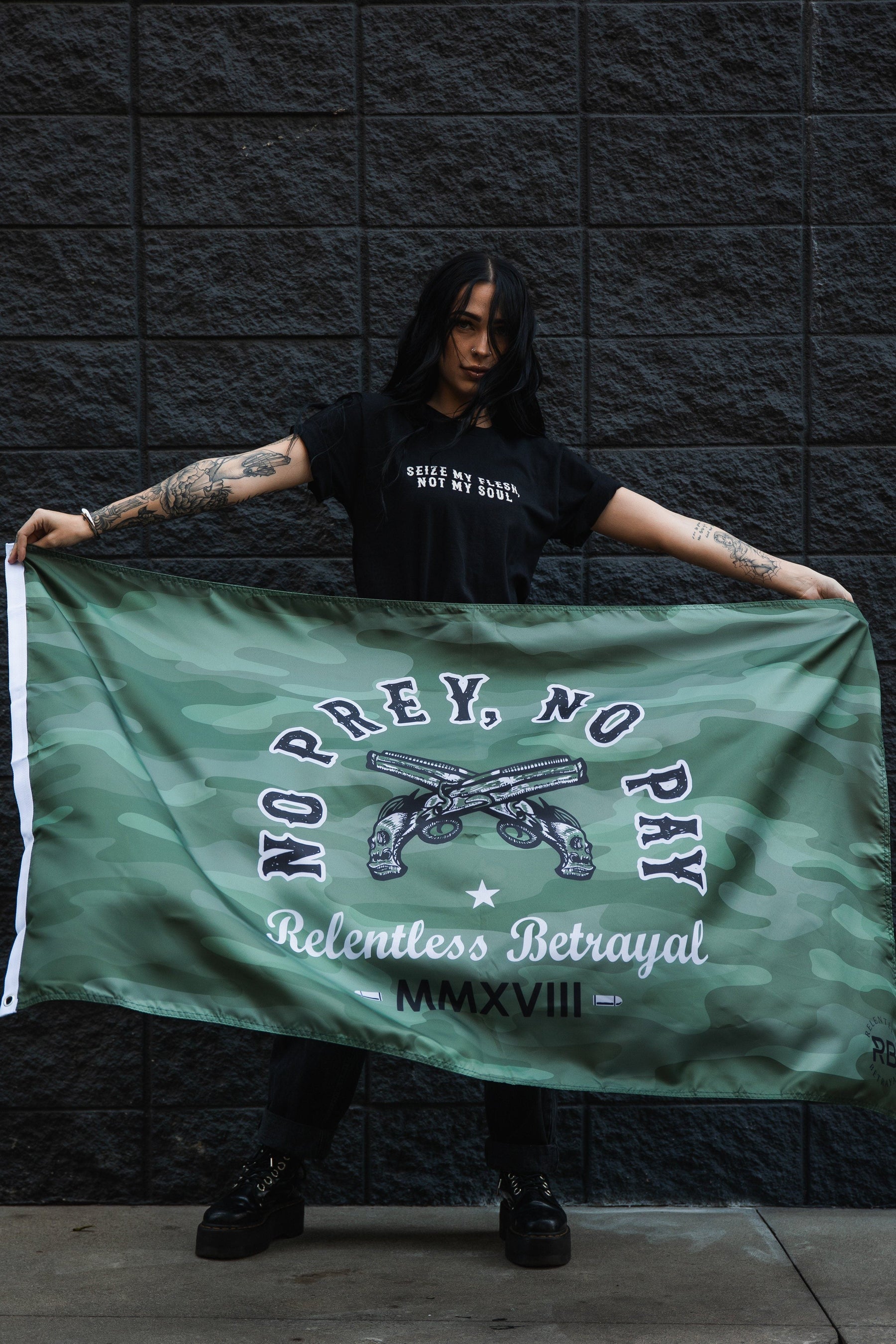 A tattooed person holds the No Prey, No Pay camo flag by Relentless Betrayal, made of durable polyester and featuring MMXVII. They stand against a dark brick wall in a black t-shirt with white text and matching pants.