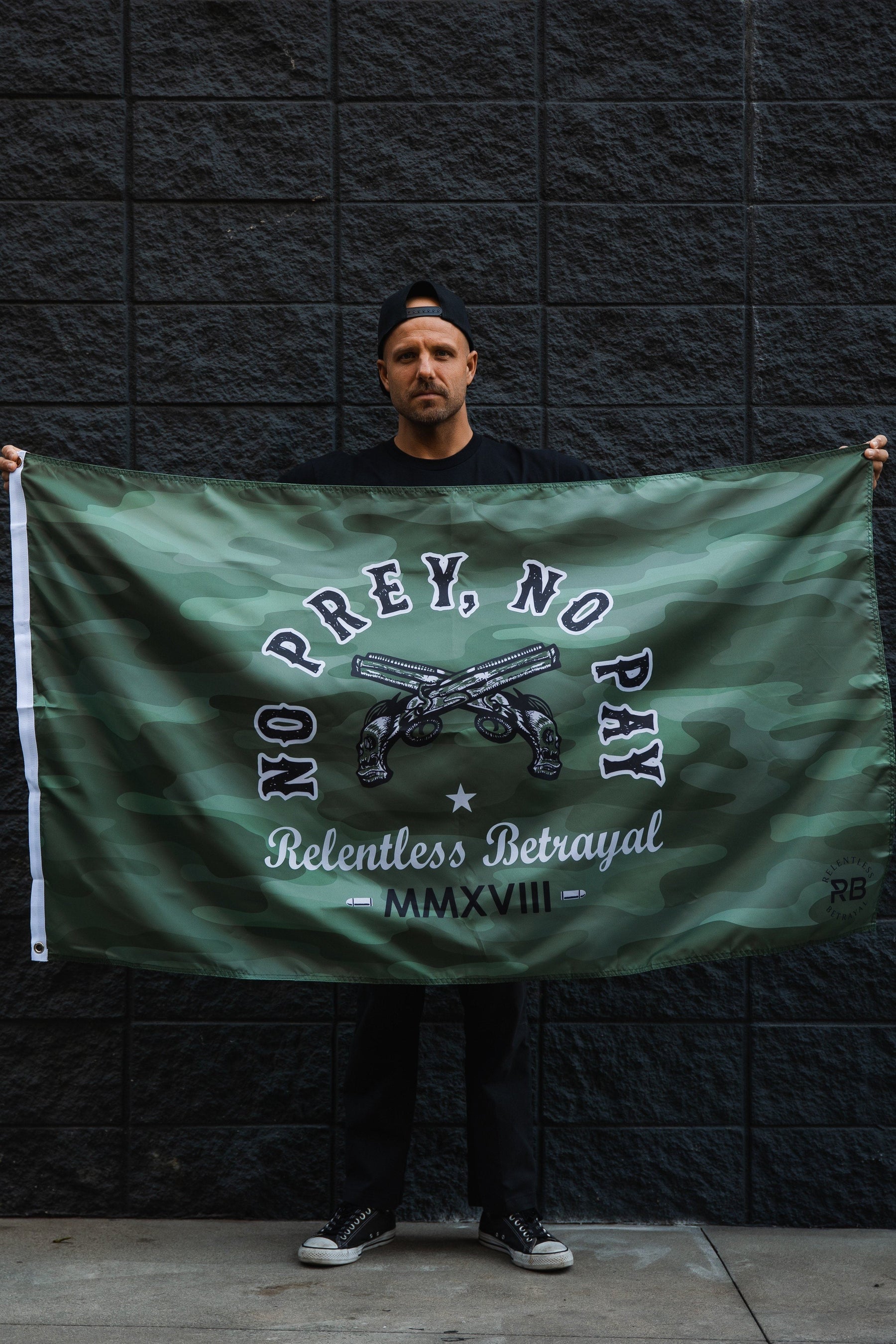 A person stands against a dark brick wall, holding the Relentless Betrayals No Prey, No Pay Camo Flag, made of durable polyester with crossed guns, a star, and the words Relentless Betrayal and MMXVIII. They sport a backward cap and dark attire.