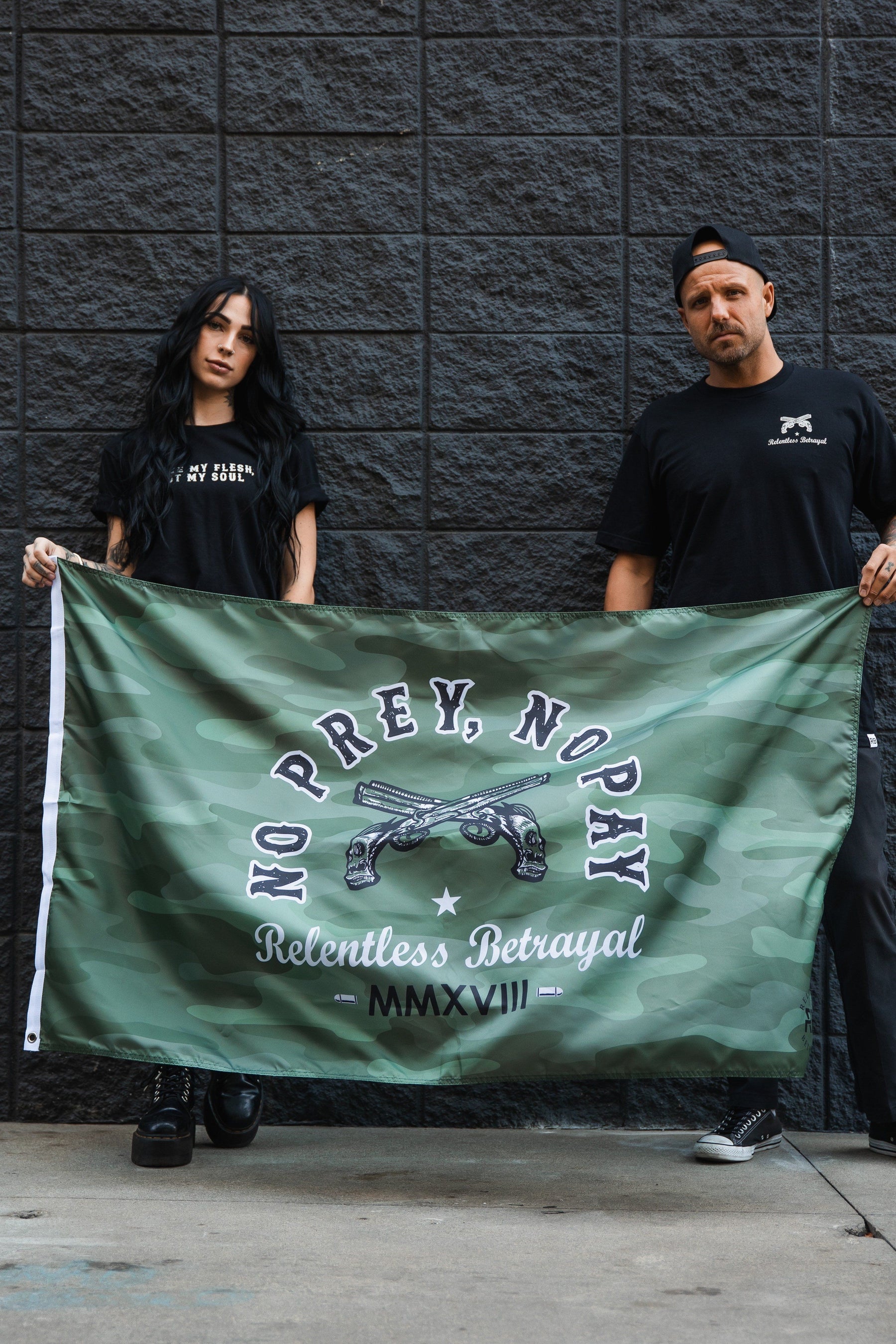 Two people, dressed in black with serious expressions, stand against a gray brick wall holding the durable polyester No Prey, No Pay Camo Flag by Relentless Betrayal featuring crossed revolvers and the text NO PREY, NO PAY - Relentless Betrayal - MMXVII.
