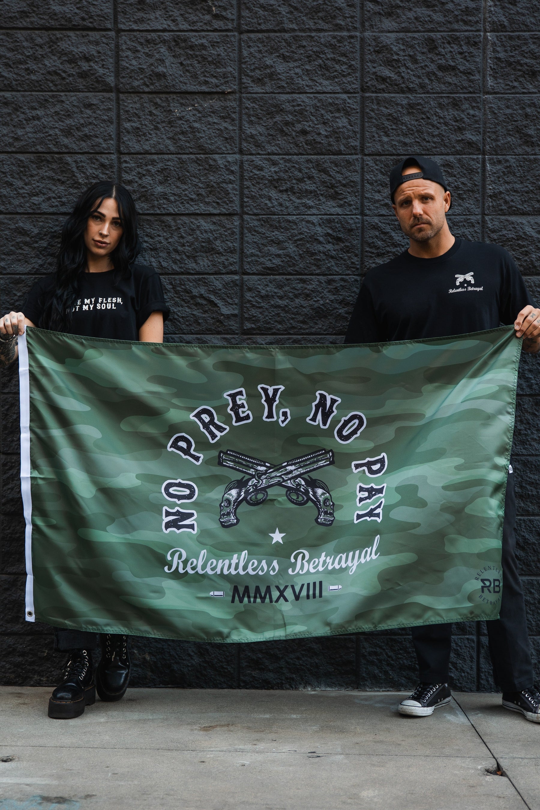 In front of a dark brick wall, two people hold the No Prey, No Pay Camo Flag by Relentless Betrayal, featuring crossed pistols and text about relentless betrayal. Their black attire and serious expressions perfectly match the rebellious spirit of the polyester flag displaying No Prey, No Pay.