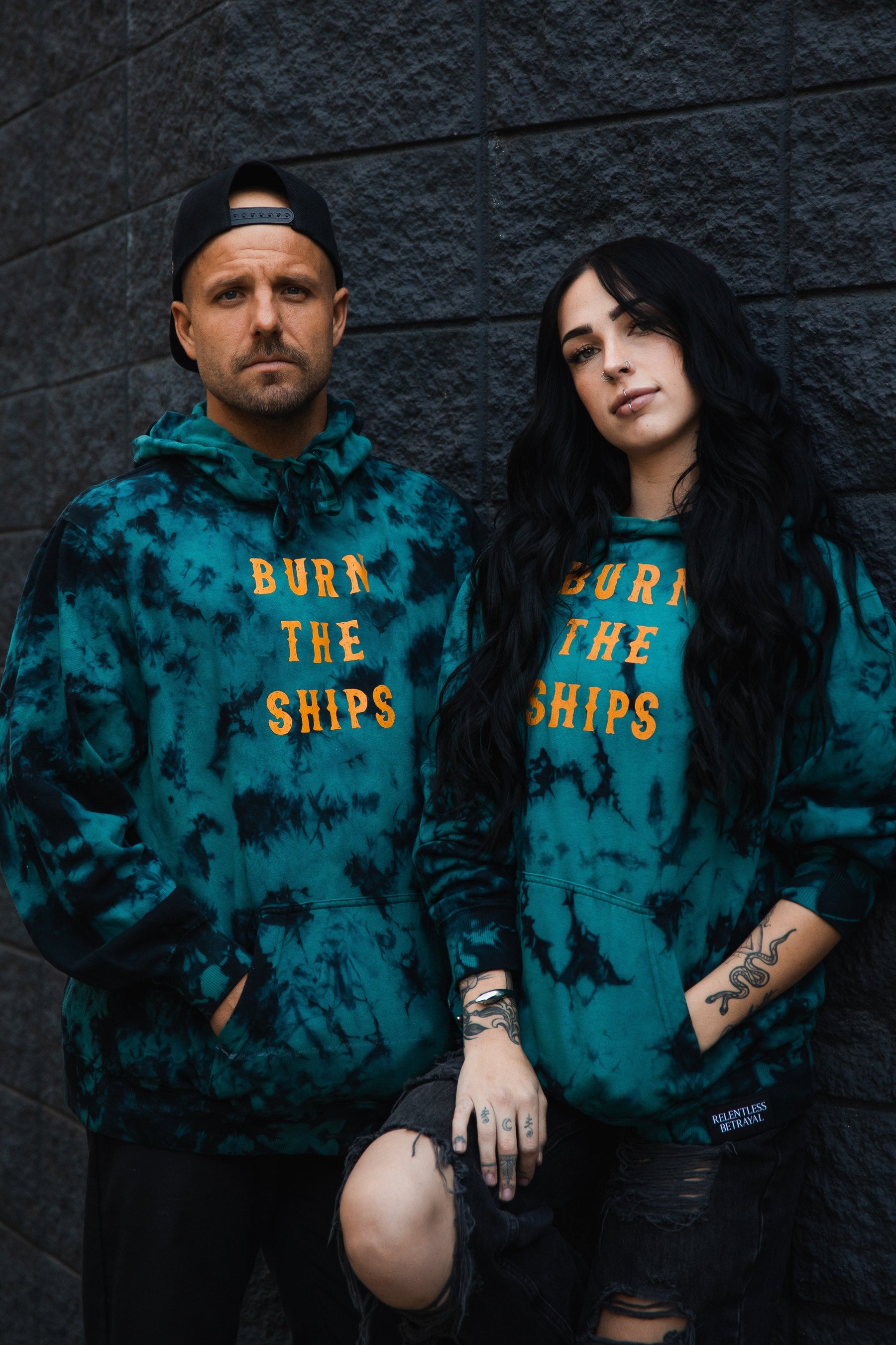 Two people lean against a dark brick wall in matching Relentless Betrayal Burn The Ships Tie Dye Hoodies, crafted from a cozy cotton-polyester blend. The teal unisex hoodies feature bold yellow text. One person wears a backward cap; the other sports long black hair and a tattooed arm.