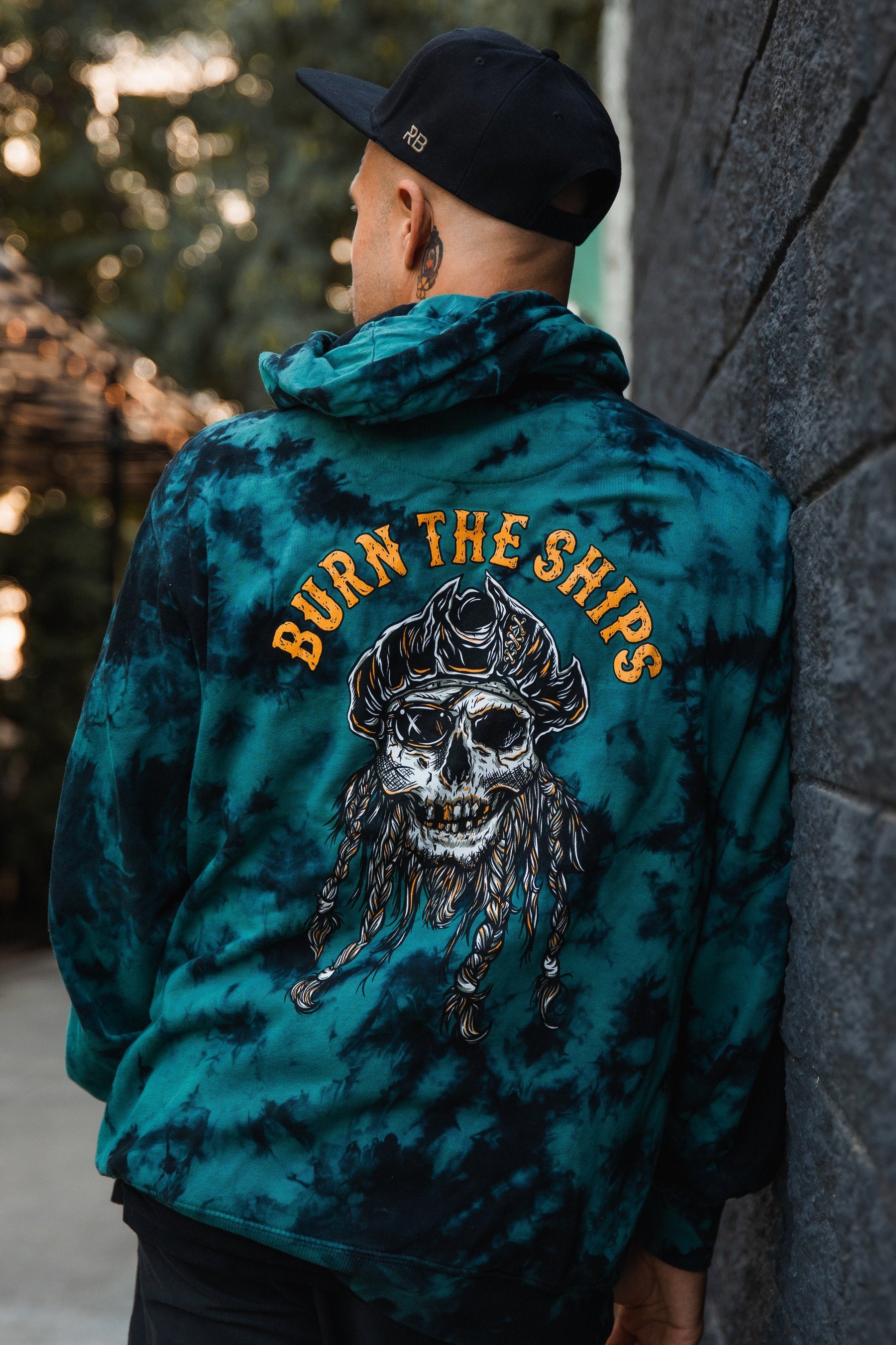 Wearing a Burn The Ships tie-dye hoodie by Relentless Betrayal, featuring a skull design on the back, a man stands against a dark wall with an outdoor blur in the background. Crafted from a cotton-polyester blend for unisex appeal.