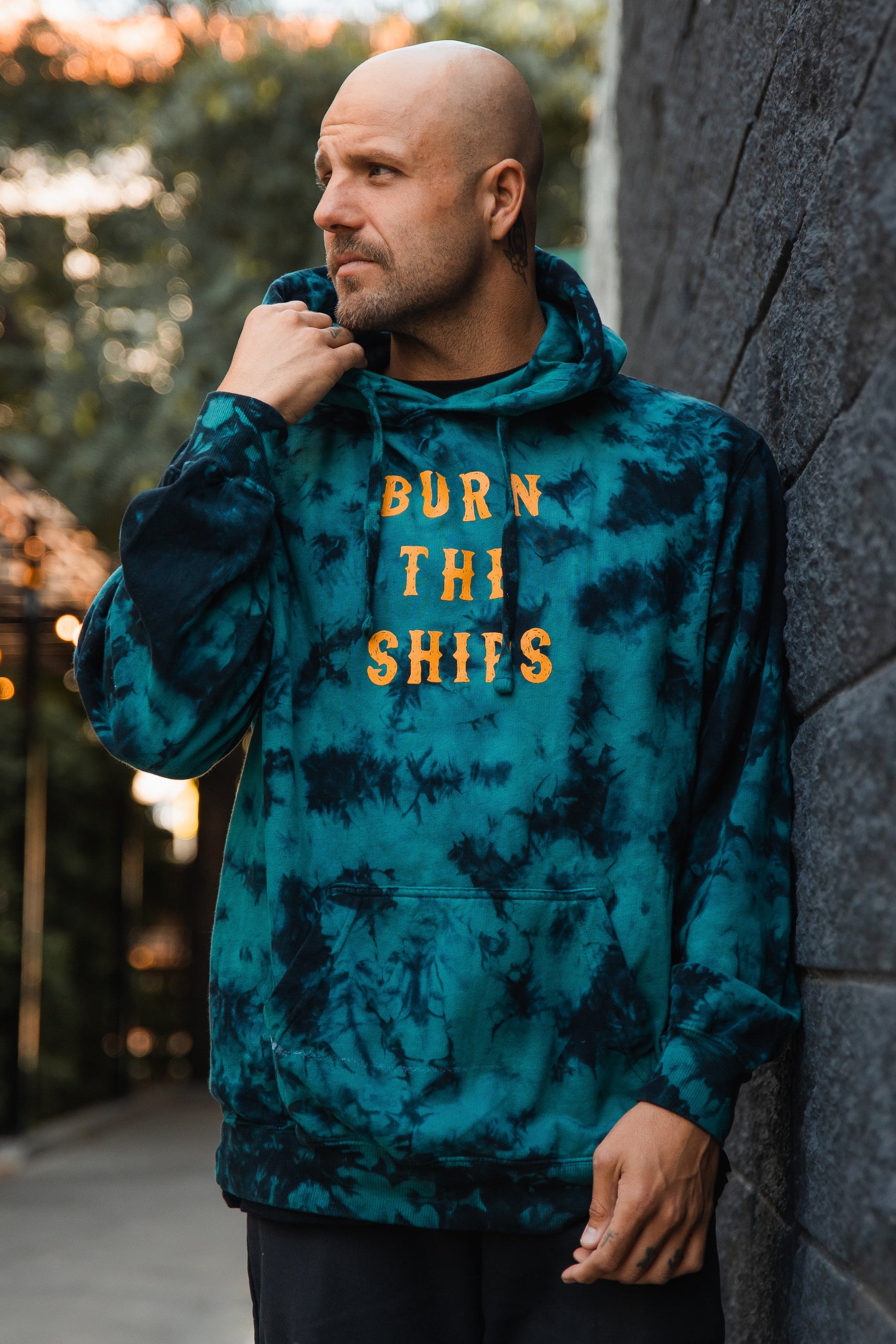 A bald man stands outdoors near a dark stone wall, gazing thoughtfully to the side. He wears a Relentless Betrayal Burn The Ships tie-dye hoodie in a blue cotton polyester blend, featuring bold orange letters.