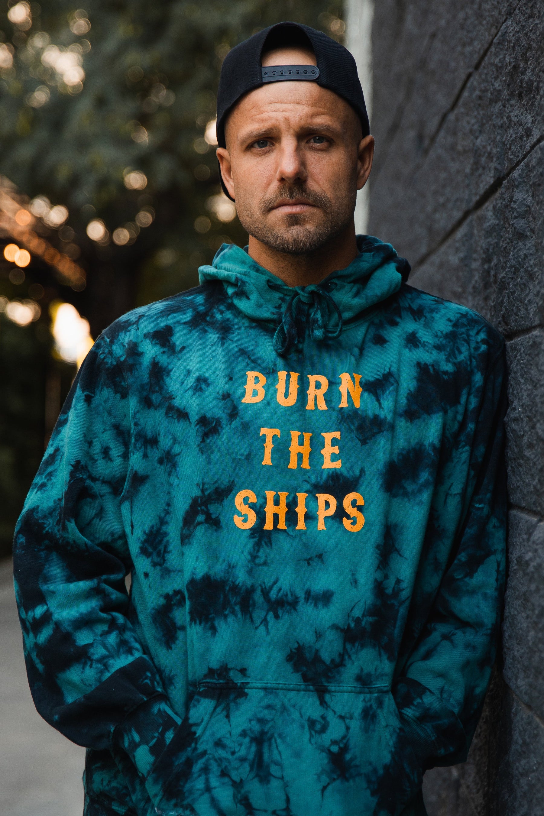A man poses against a textured wall in a teal tie-dye unisex hoodie from Relentless Betrayal, featuring Burn The Ships in yellow. His serious look matches his backwards black cap, with trees and a path subtly blurred in the background.