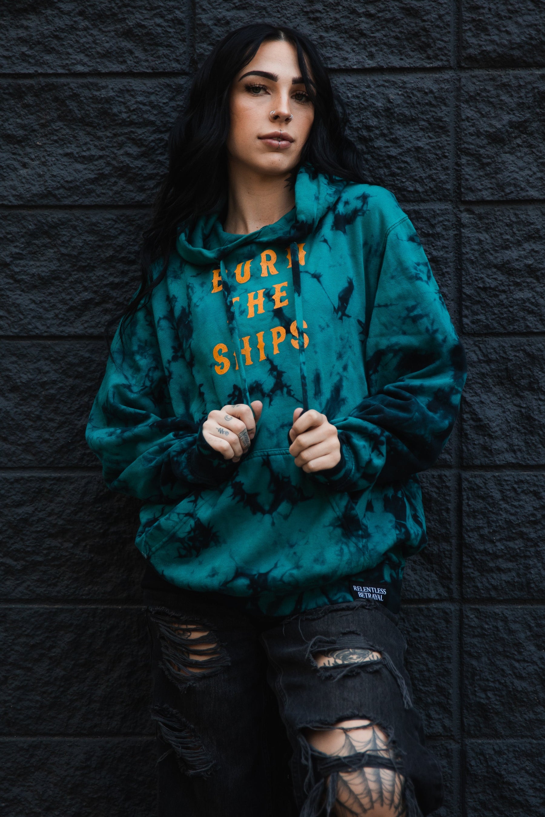 A person with long dark hair stands against a textured dark wall, wearing a Relentless Betrayal Burn The Ships Tie Dye Hoodie made from a cotton polyester blend and ripped black jeans. They have a relaxed expression and casually grip the hoodie near the collar.