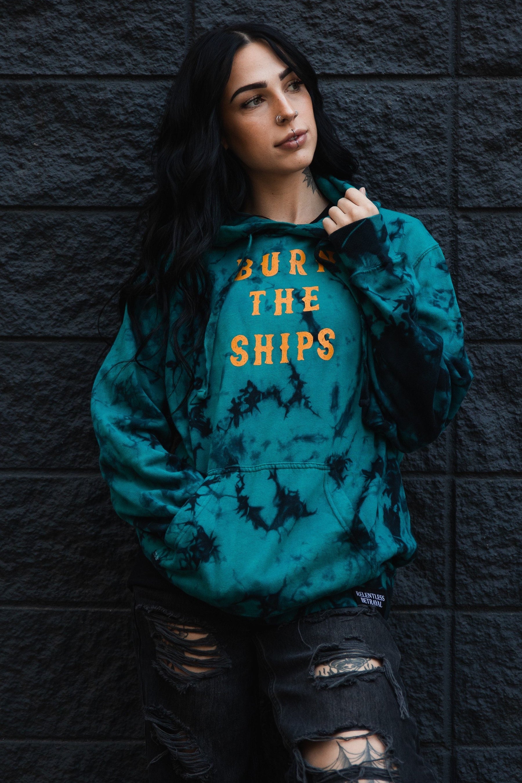 A woman with long dark hair stands against a textured dark wall, wearing the Burn The Ships Tie Dye Hoodie from Relentless Betrayal. The teal cotton-polyester blend features BURN THE SHIPS in yellow. Shes paired it with ripped black jeans, holding the hoodie collar and showcasing her pierced nose.